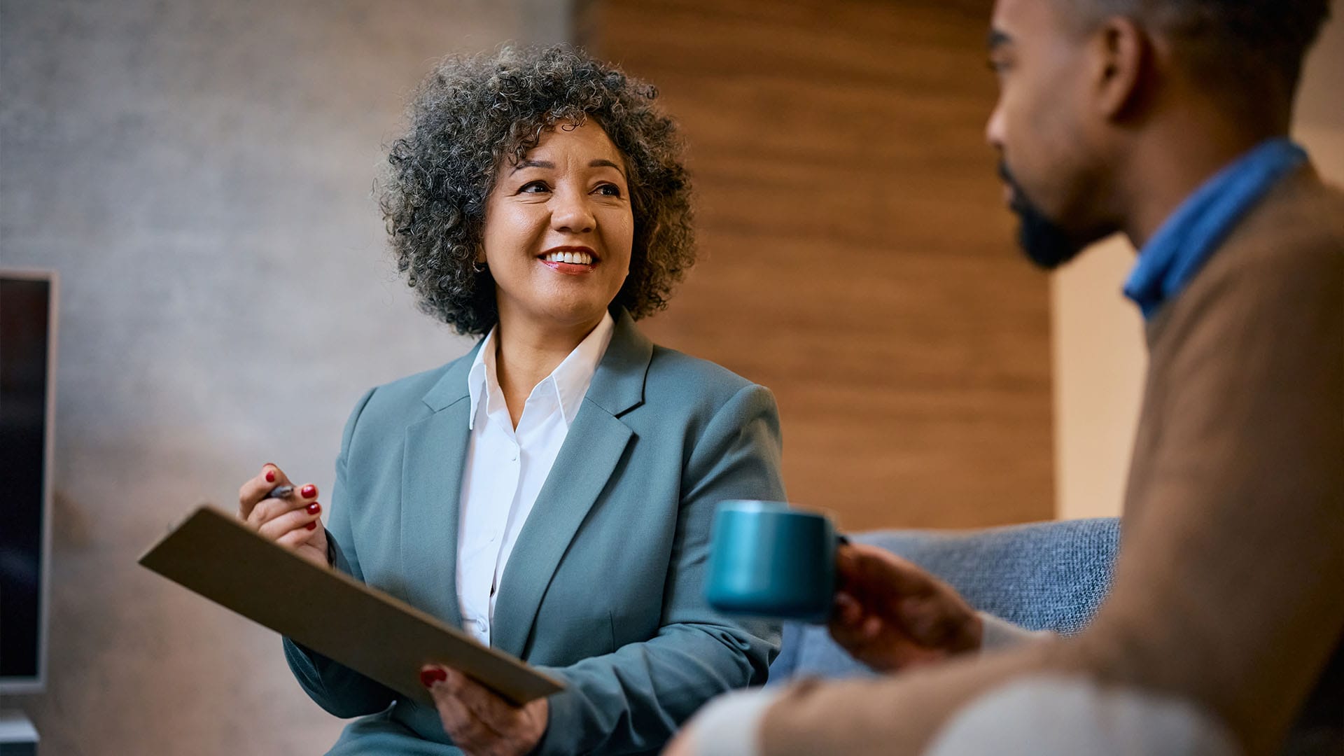 Smiling therapist holding a clipboard, engaged in conversation with a client holding a teal mug.