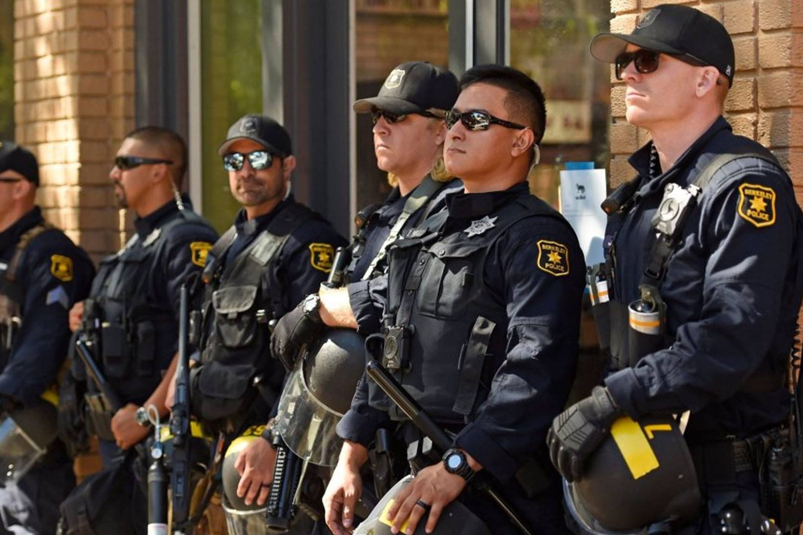 Berkeley police officers in tactical uniforms lined up against a brick wall holding batons and riot gear.