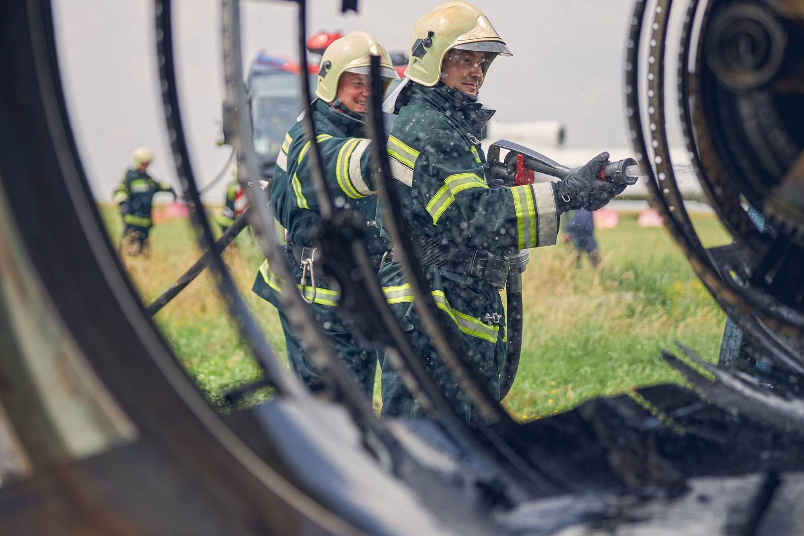 Two firefighters in yellow helmets and black protective suits are seen through the metal wreckage of a large industrial structure, working together with a hose to spray water, demonstrating teamwork during an intense emergency response.