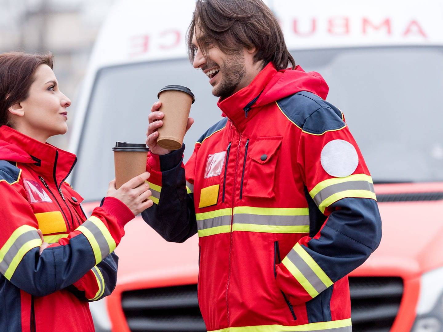Two paramedics smiling and sharing coffee outside ambulance.