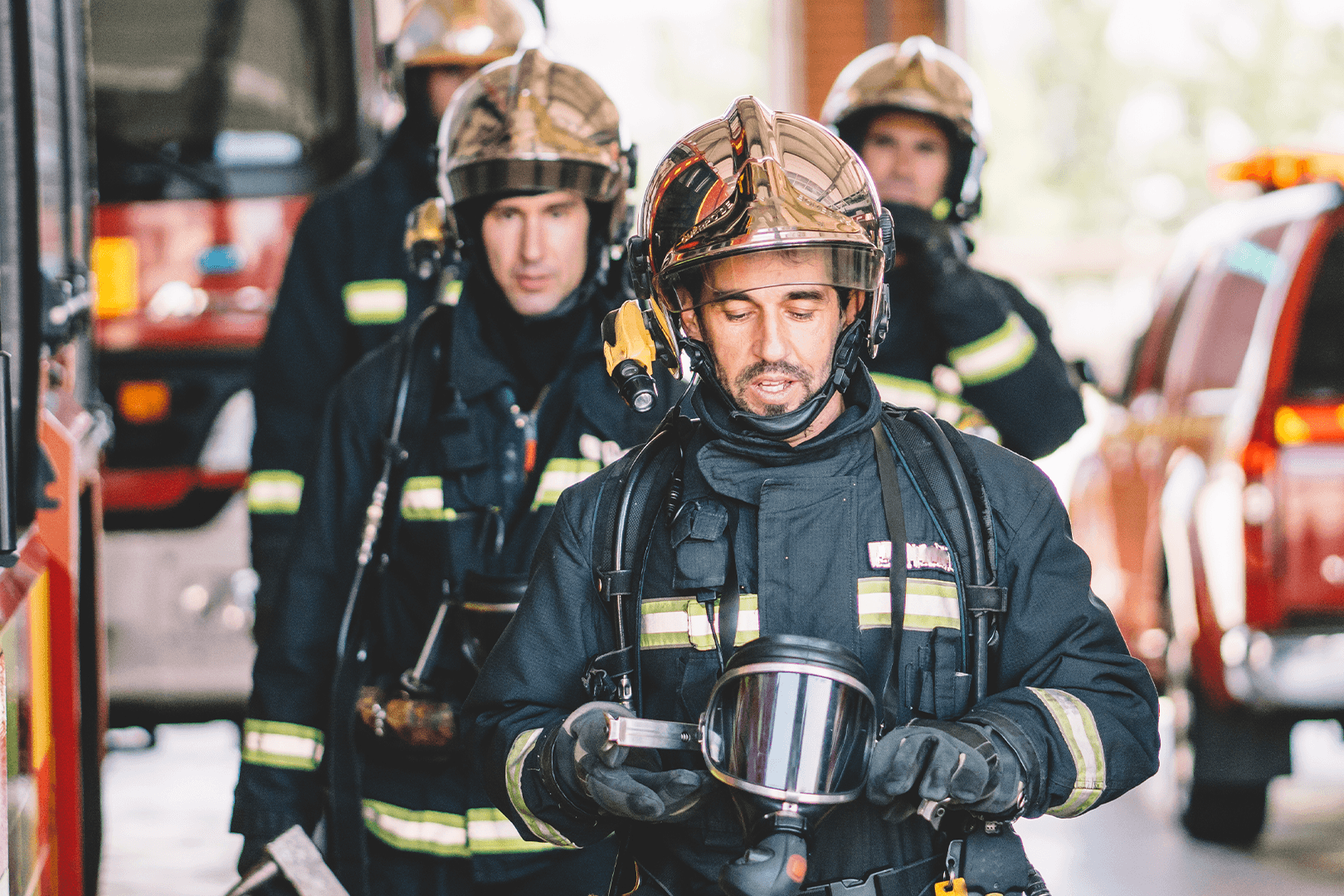 A group of Pleasanton firefighters in full protective gear walk between fire trucks, with the lead firefighter holding his helmet and looking down thoughtfully.