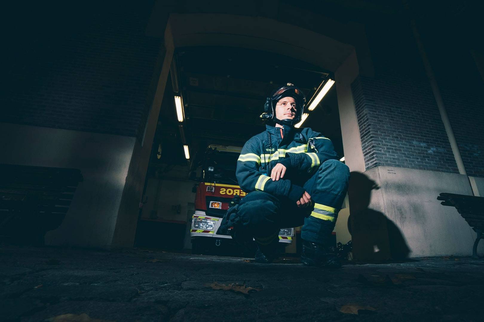 Firefighter in full gear kneeling outside a dimly lit station at night, looking contemplative.