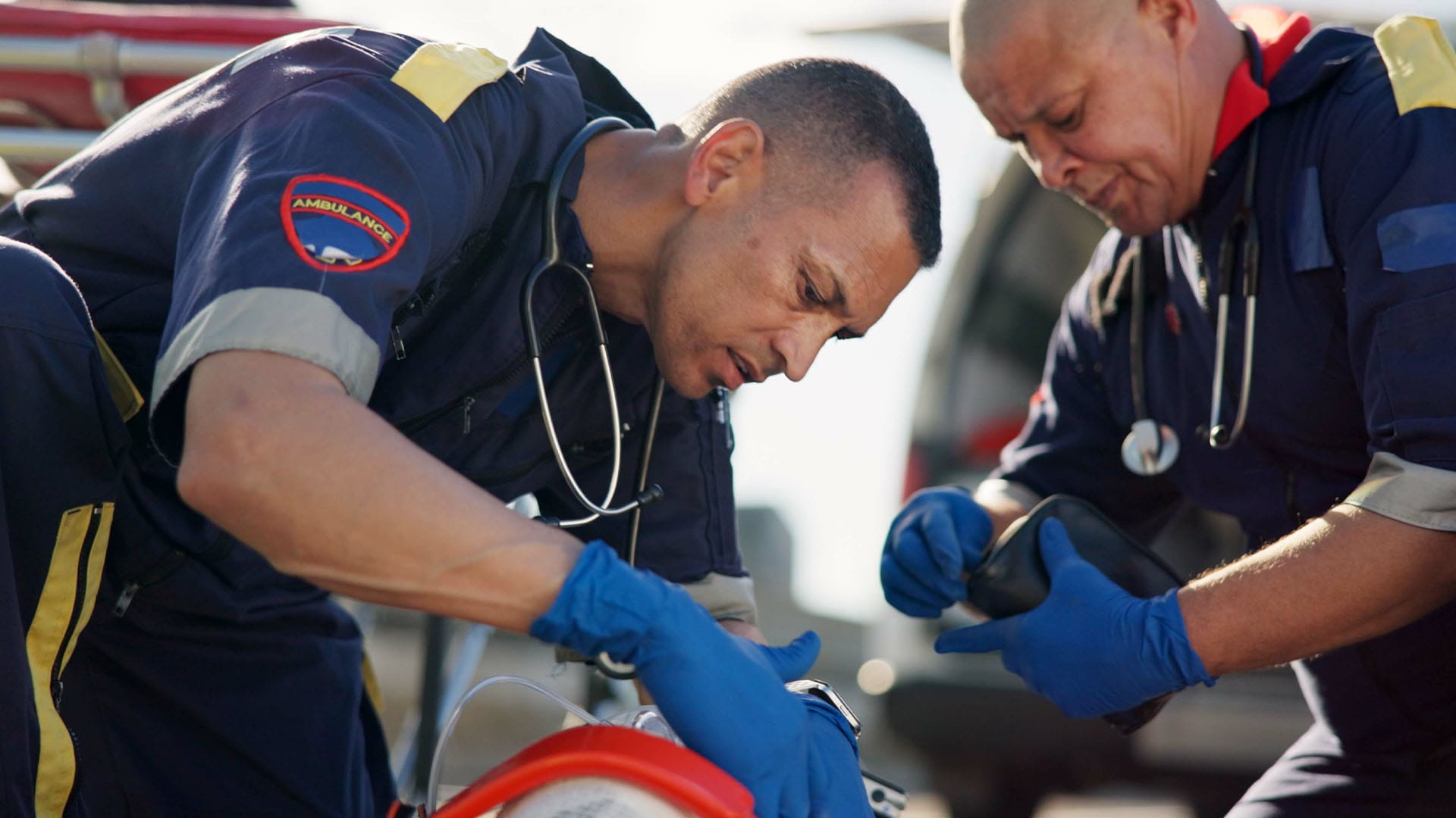 Two emergency medical technicians providing intense roadside care to a patient while wearing navy uniforms and blue gloves.