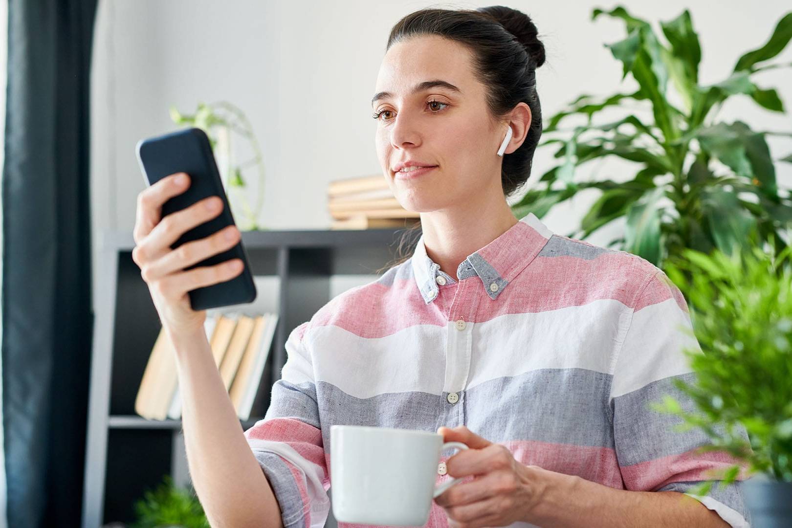 Woman in a striped button-up shirt holds a mug while participating in an online therapy session, seated near indoor plants in a home office space.