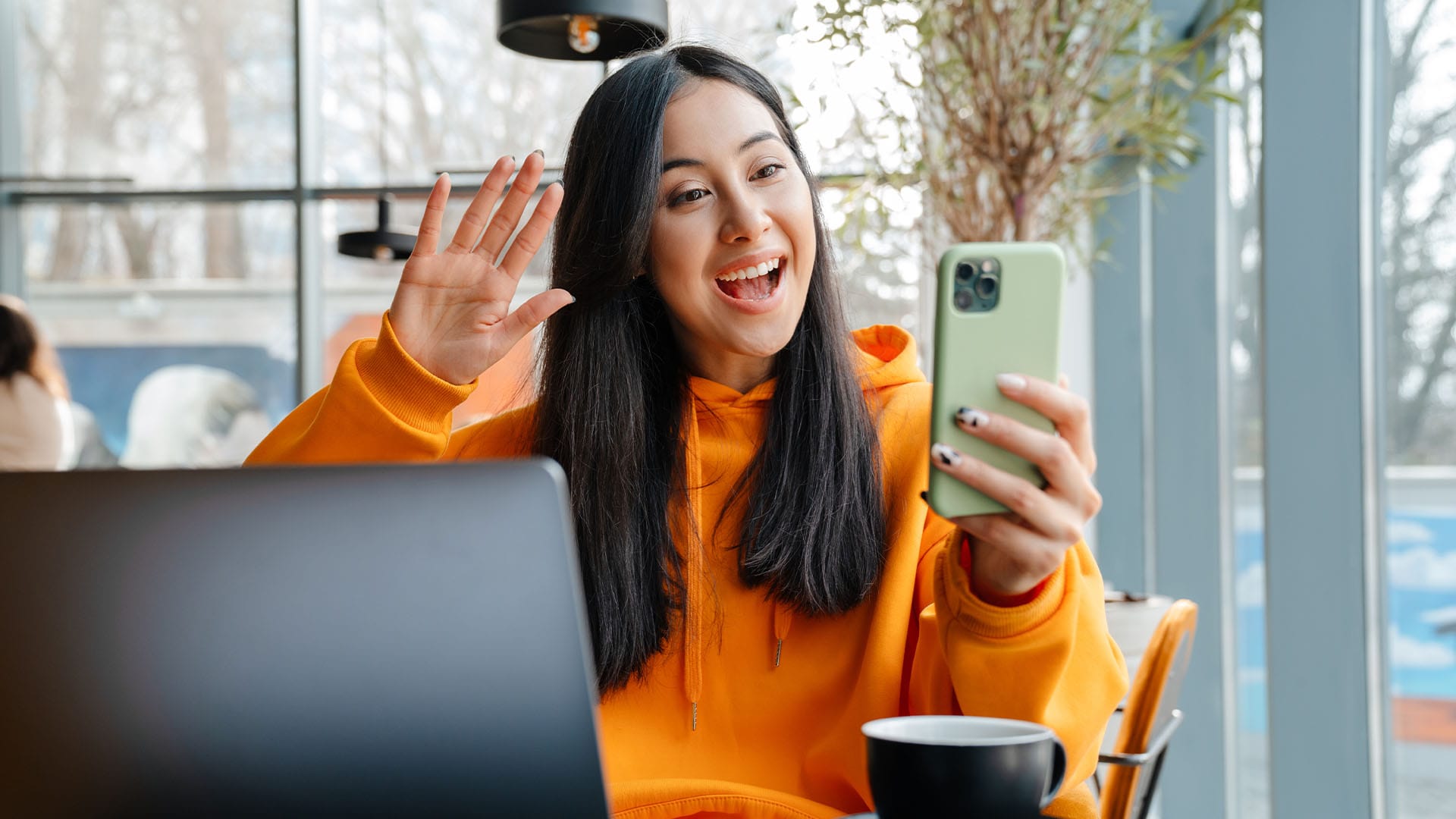 A cheerful young woman in a bright orange hoodie smiles and waves during a video call, seated at a cozy café table with a laptop and coffee mug in front of her—conveying warmth, connection, and the flexibility of remote therapy.