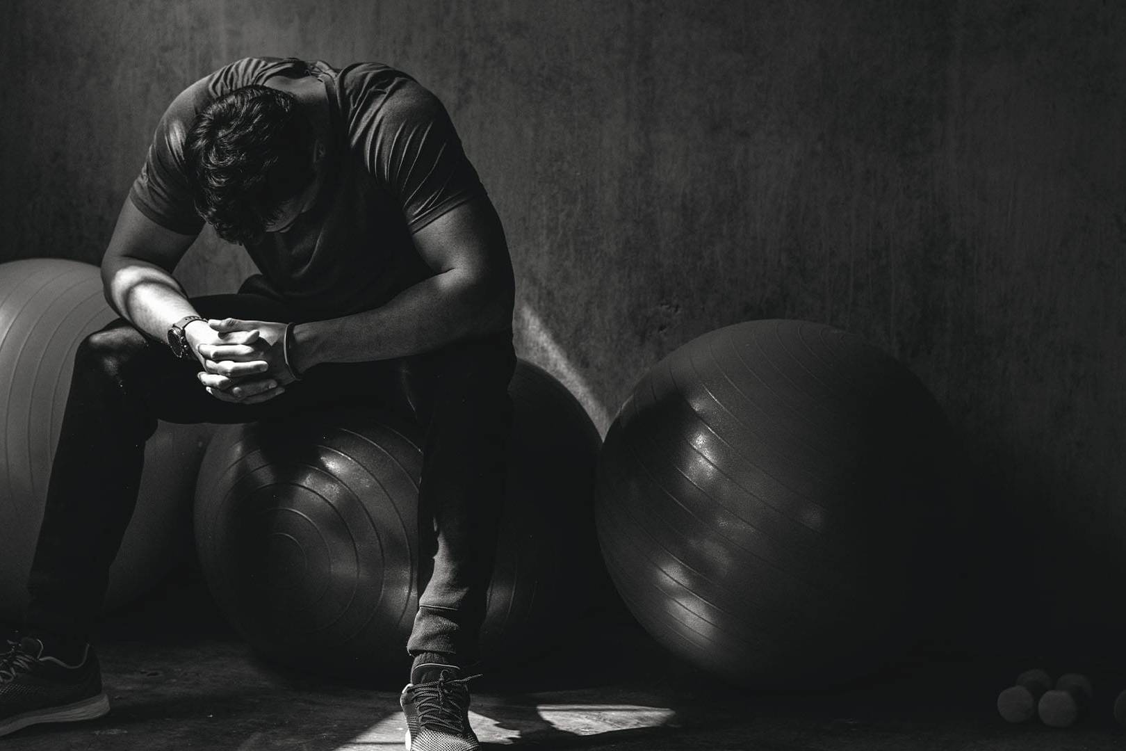 Man sitting on exercise ball in dark room with head down and hands clasped, expressing emotional distress