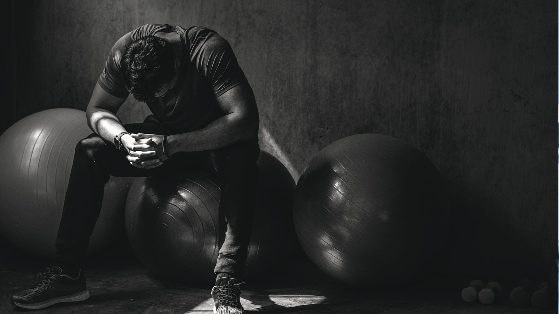 Man sitting on exercise ball in dark room with head down and hands clasped, expressing emotional distress