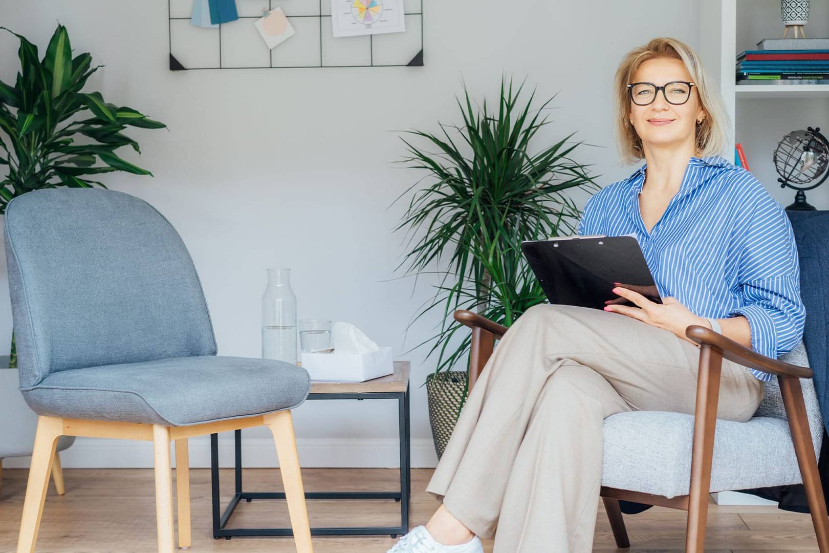 Smiling female therapist with glasses holding a clipboard while sitting in a modern office with an empty chair prepared for a client.