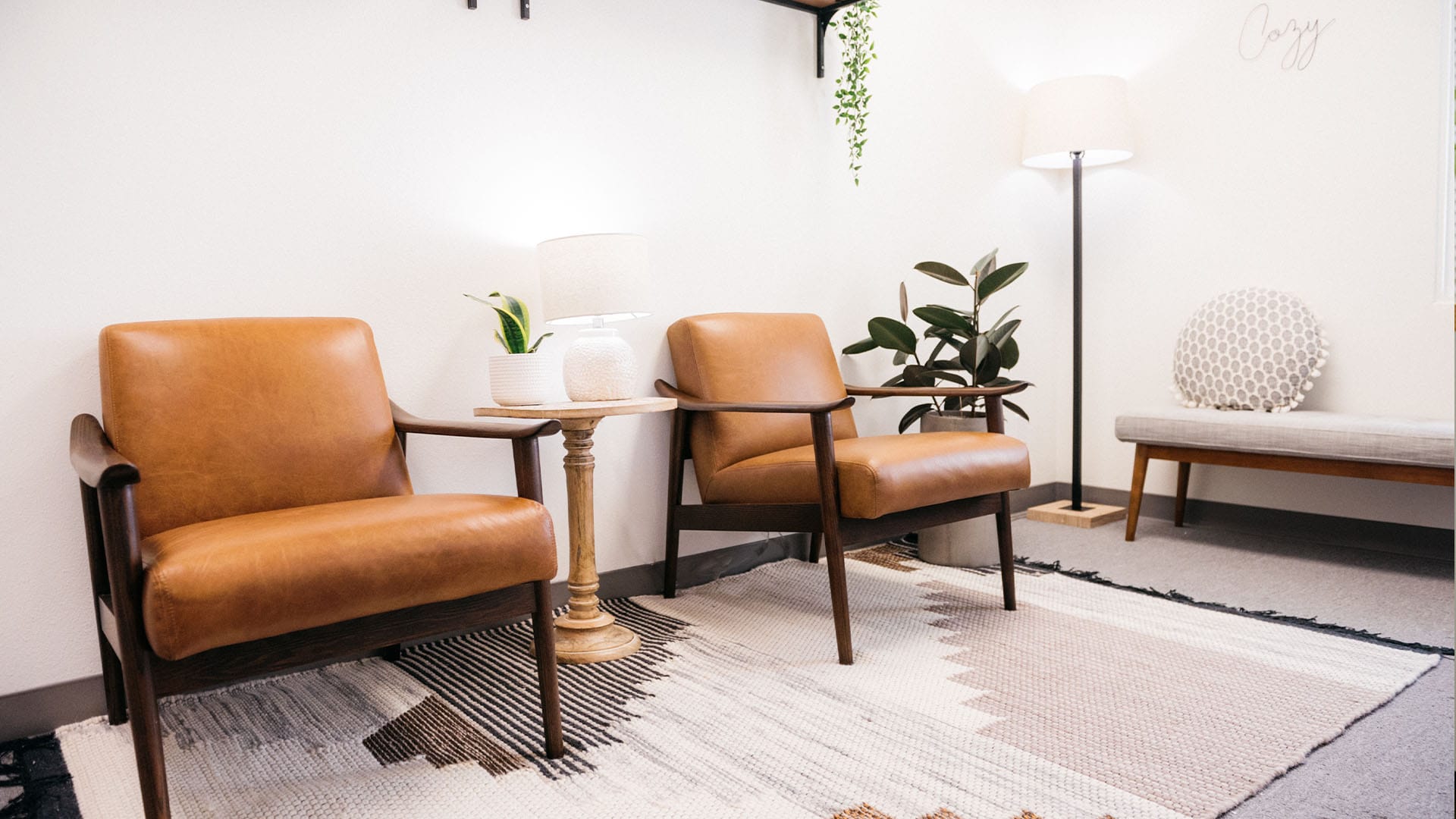 Cozy therapy office with two tan leather chairs, a floor lamp, plants, and a cushioned bench, creating a welcoming space for counseling sessions.