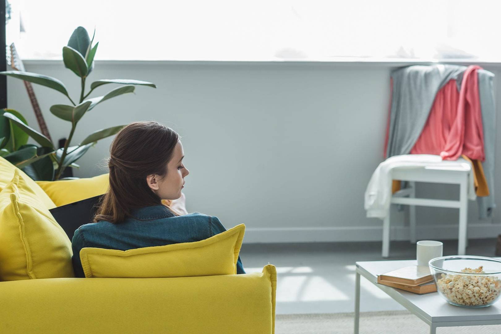 woman sitting alone on yellow couch in bright living room with popcorn on table