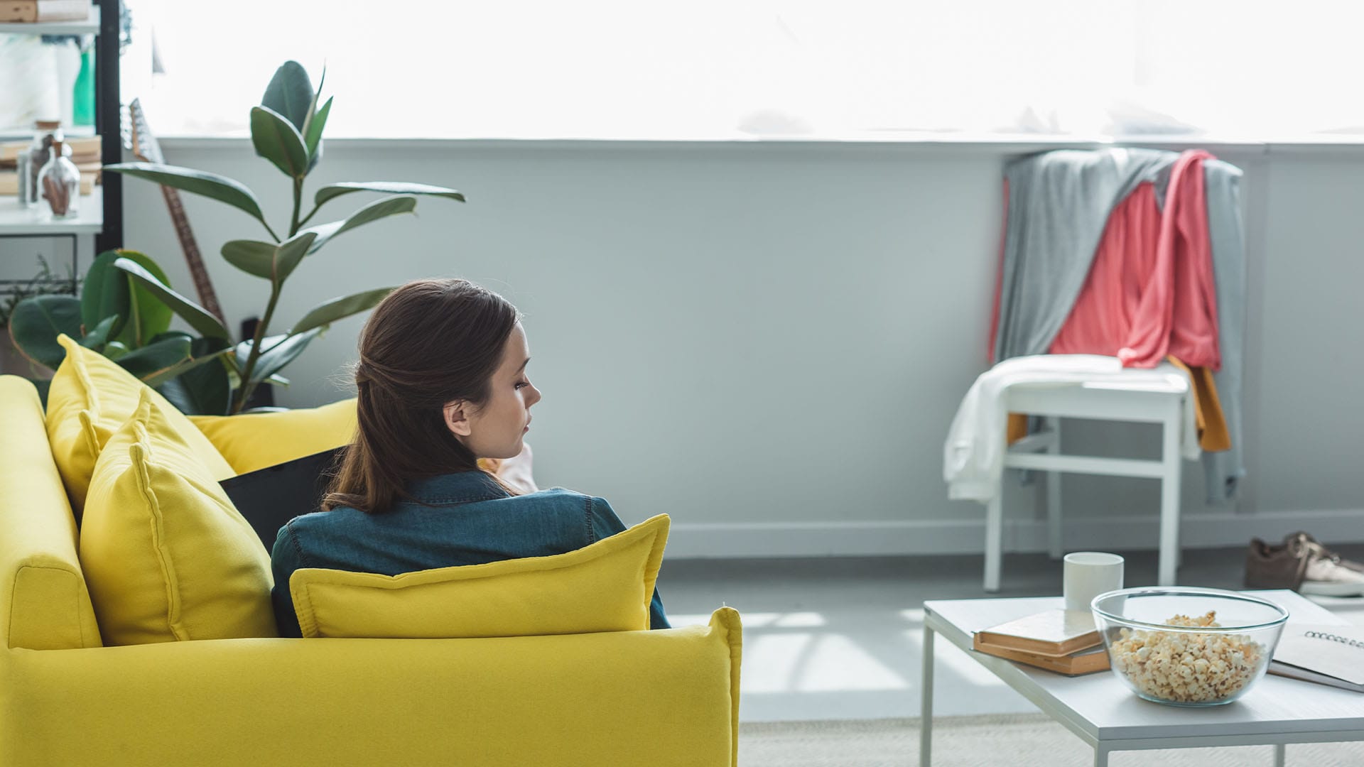 woman sitting alone on yellow couch in bright living room with popcorn on table