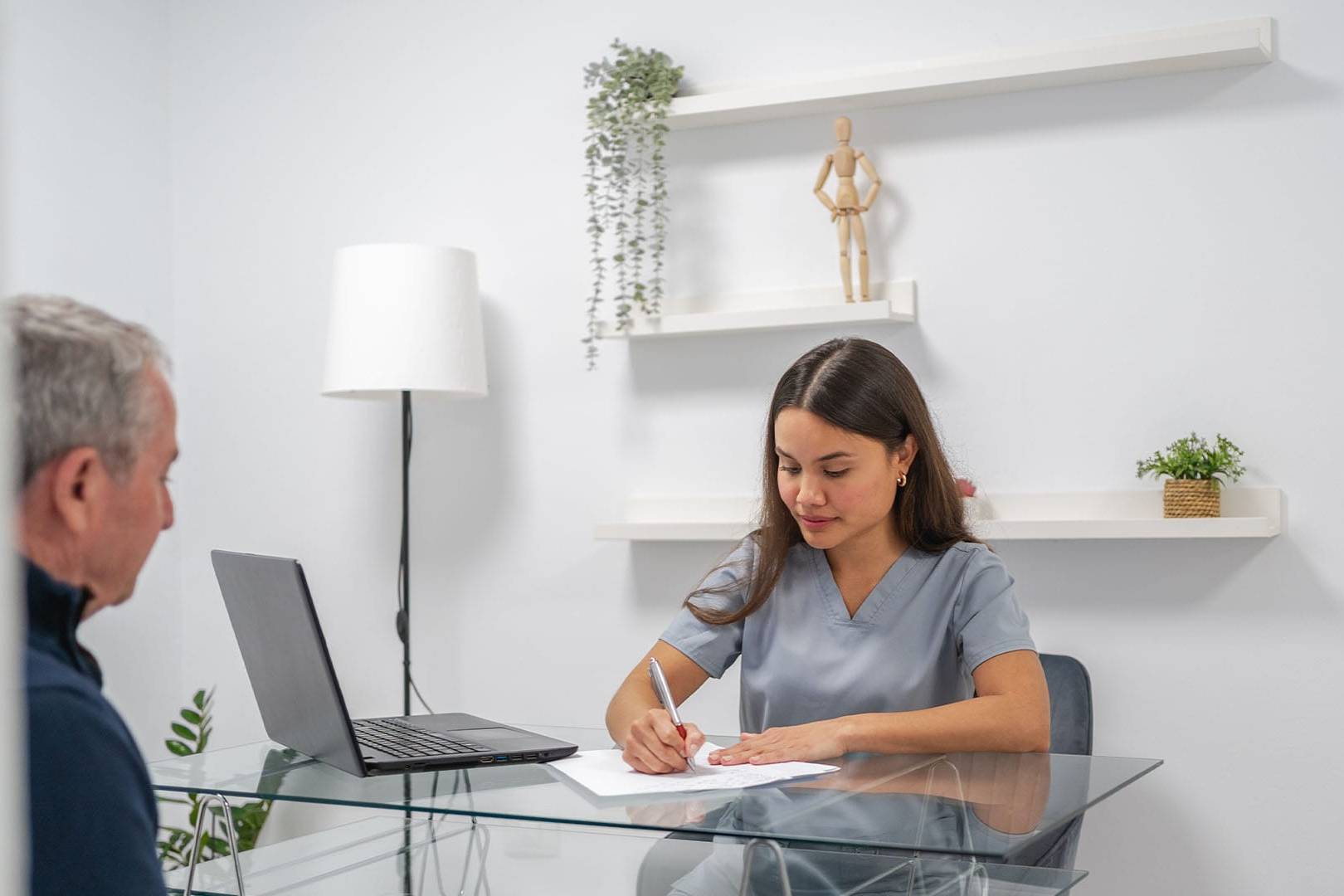 Female therapist in scrubs writing notes during a session with a male client seated across a glass desk in a modern, minimal office.