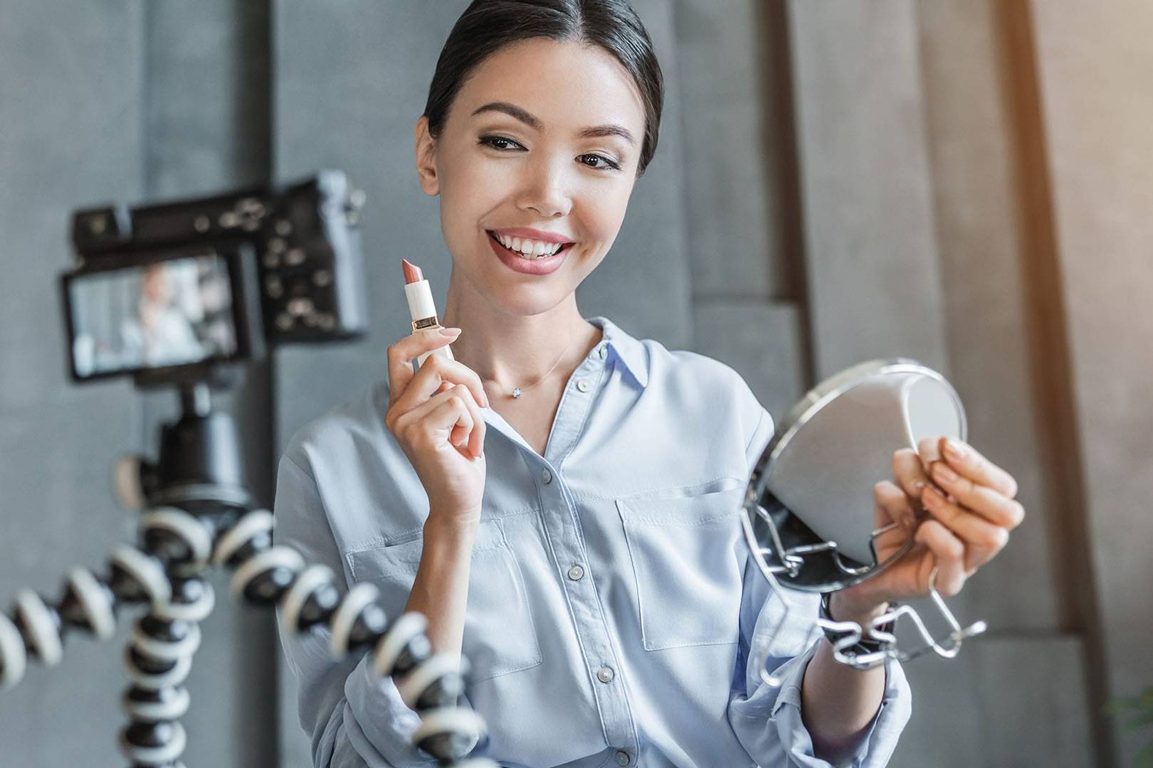 Young woman smiling while holding lipstick and mirror, filming herself on camera at home