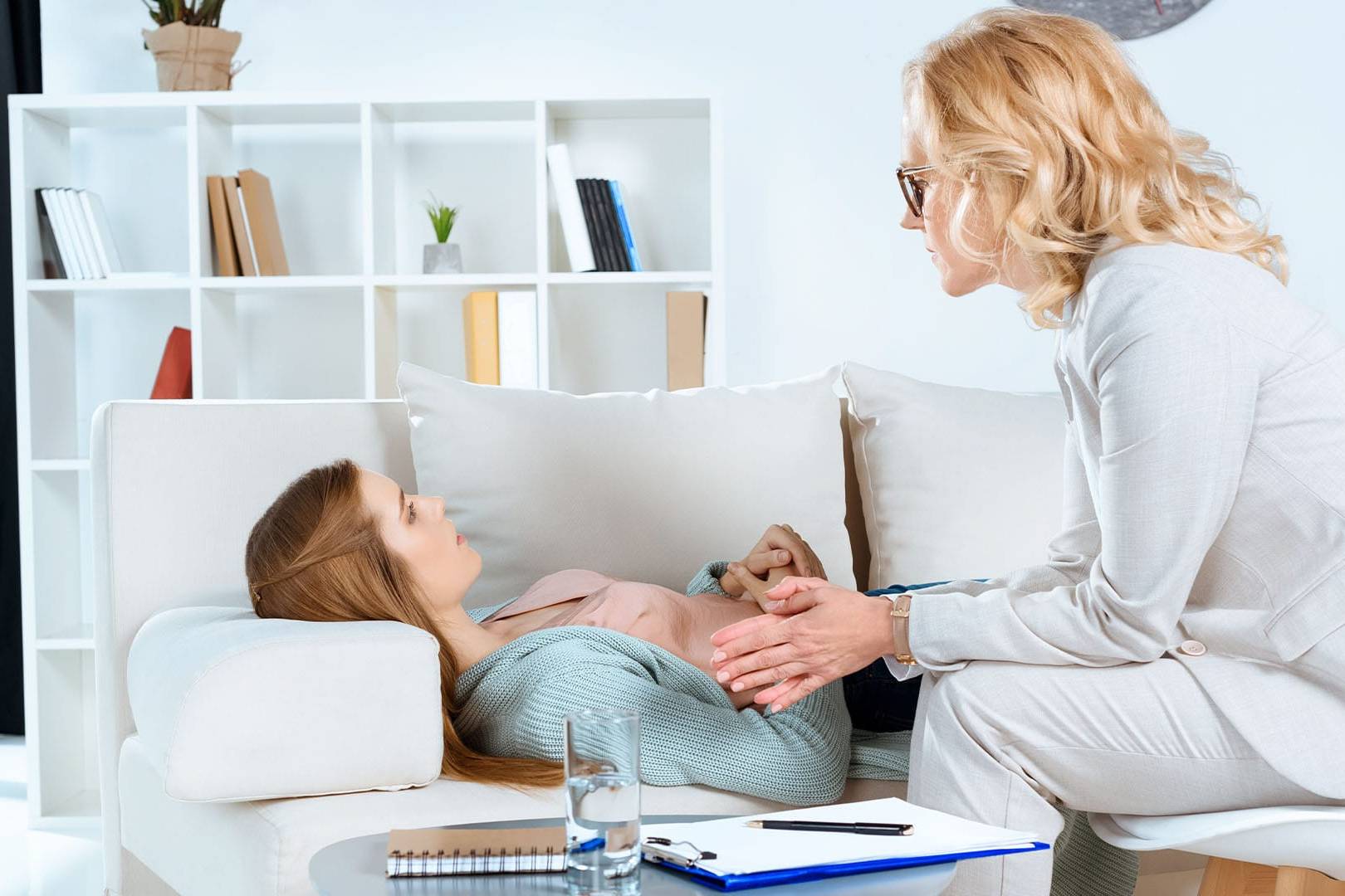 Female therapist in a light suit attentively listening to a client lying on a couch during a trauma recovery therapy session in a modern office.