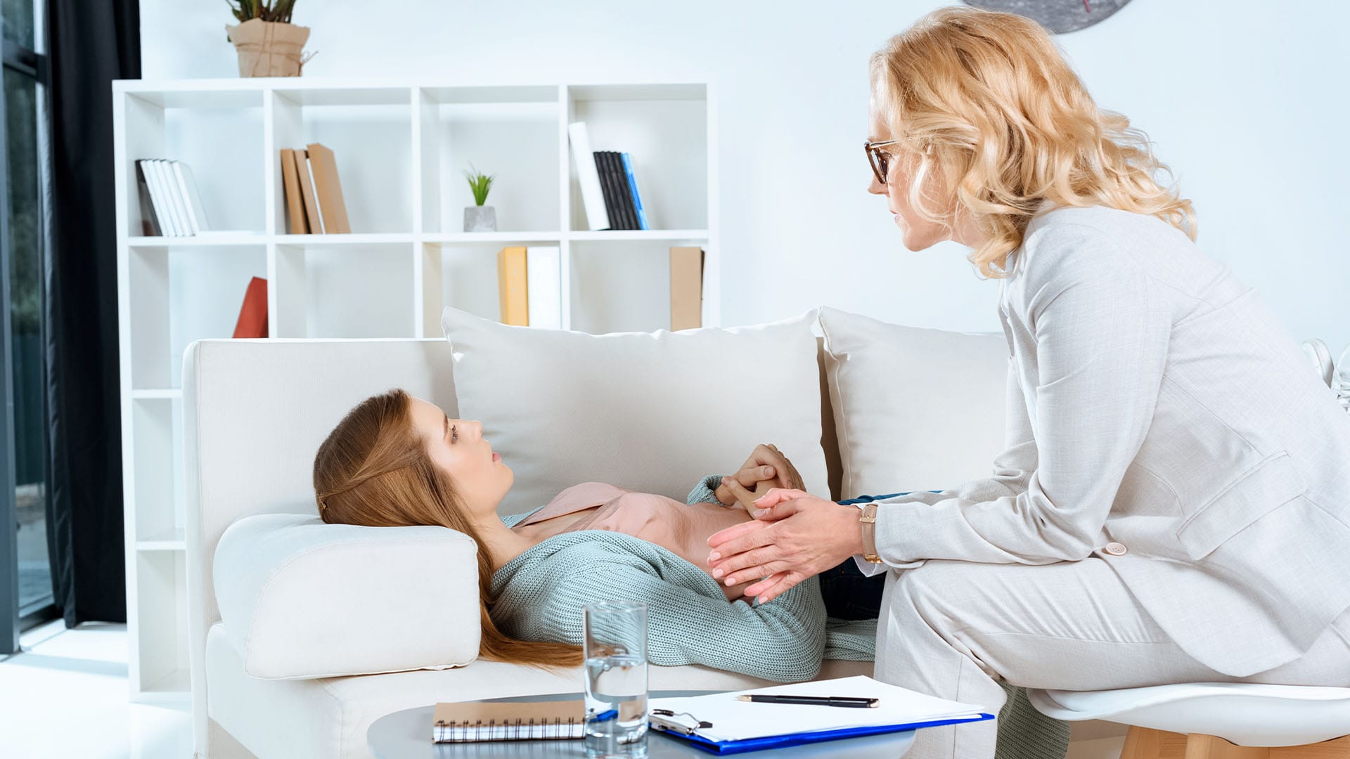 Female therapist in a light suit attentively listening to a client lying on a couch during a trauma recovery therapy session in a modern office.