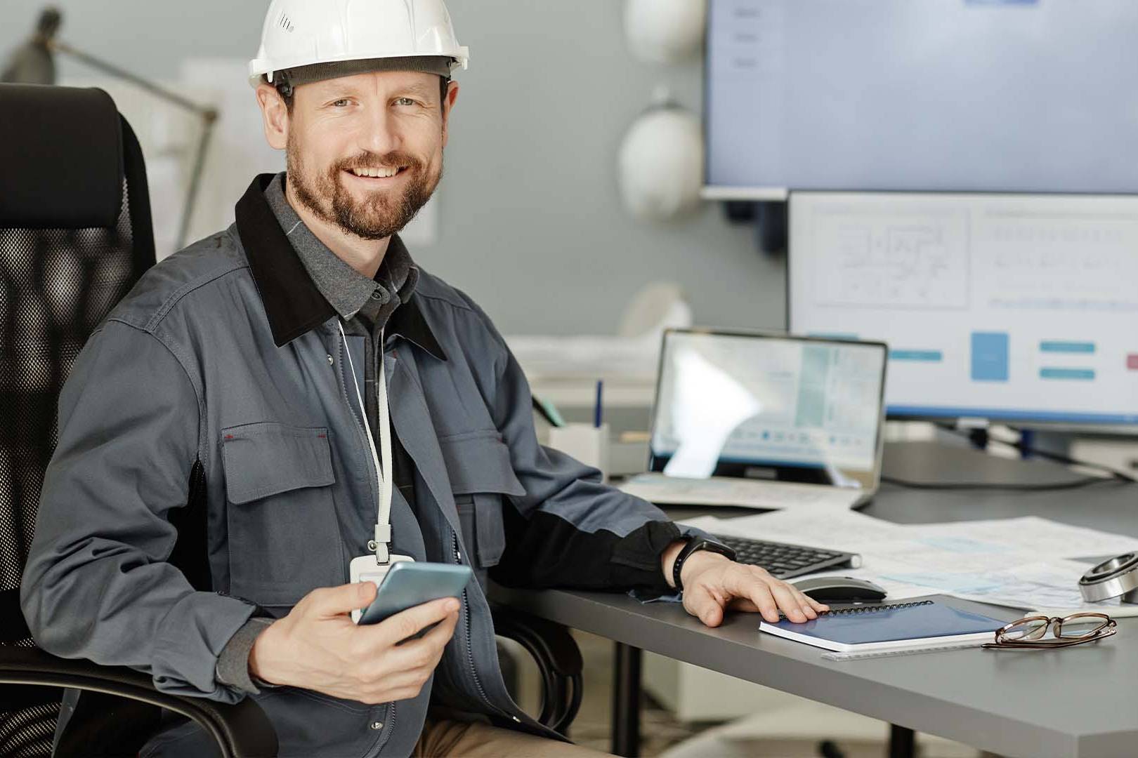Engineer in hard hat and work jacket at desk with multiple monitors holding phone in industrial office setting