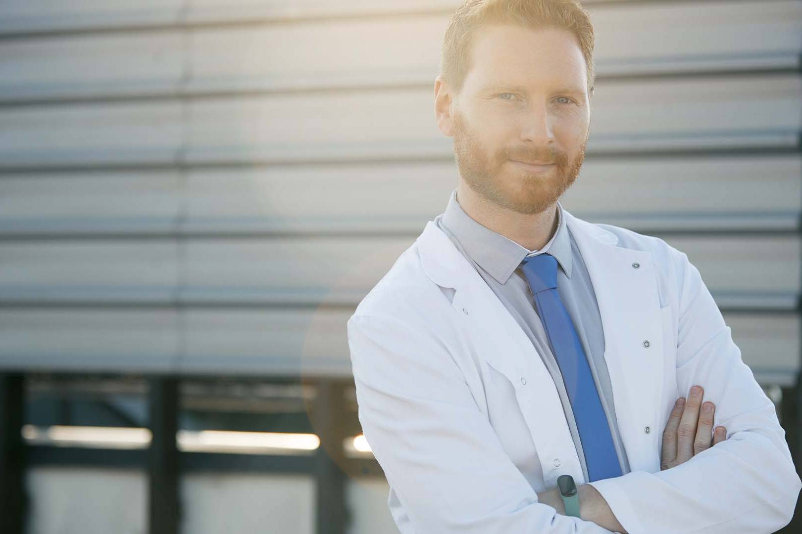 Confident male doctor in white coat standing arms crossed outdoors