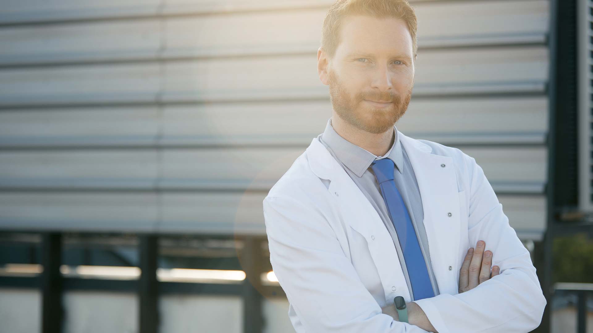 Confident male doctor in white coat standing arms crossed outdoors