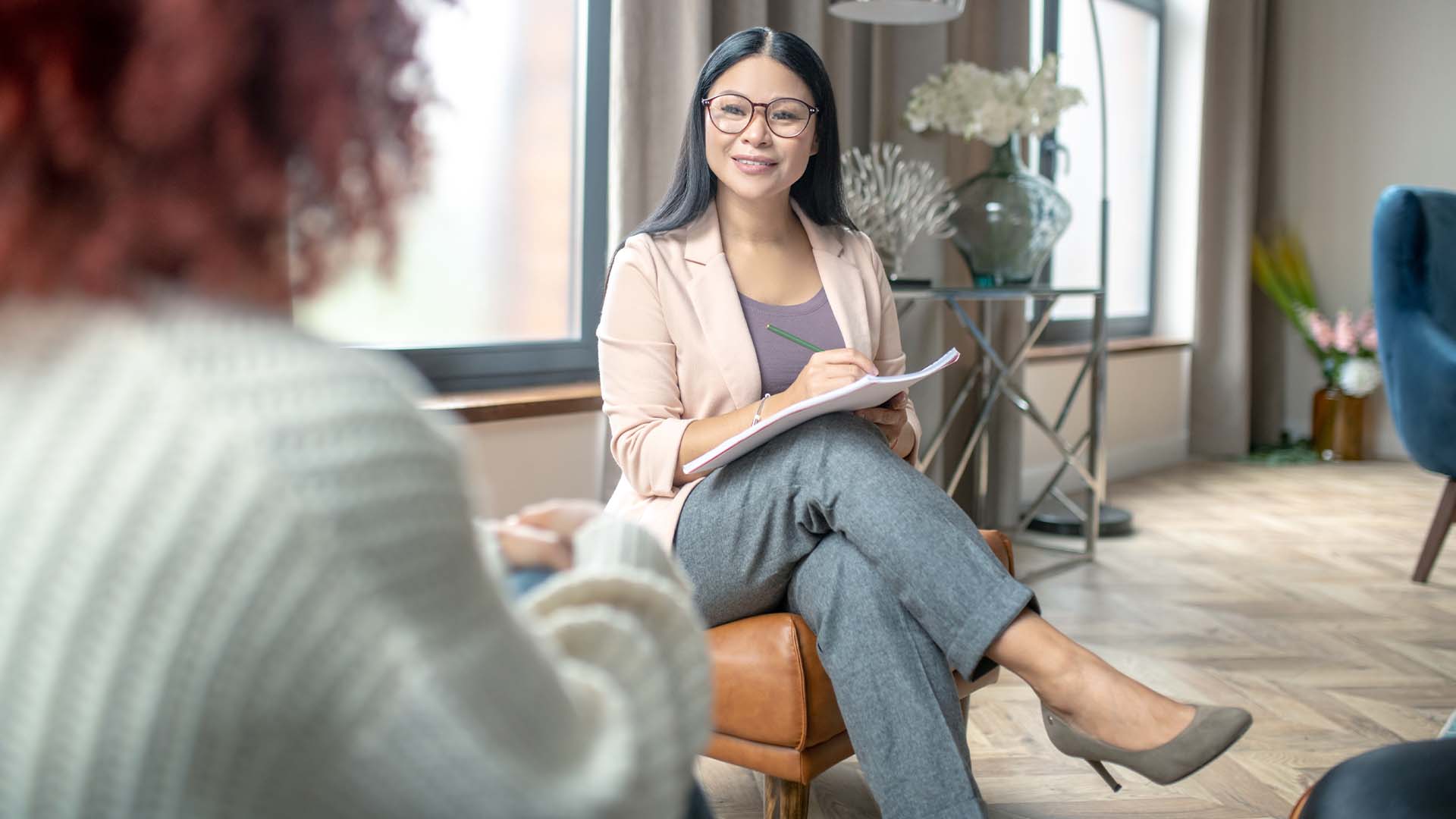 Smiling therapist with glasses taking notes during session with client in cozy office