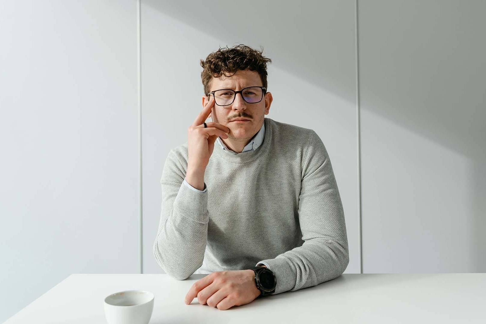 A man with curly hair and glasses sitting at a white desk with a coffee cup, resting his chin on his hand with a contemplative, exhausted expression