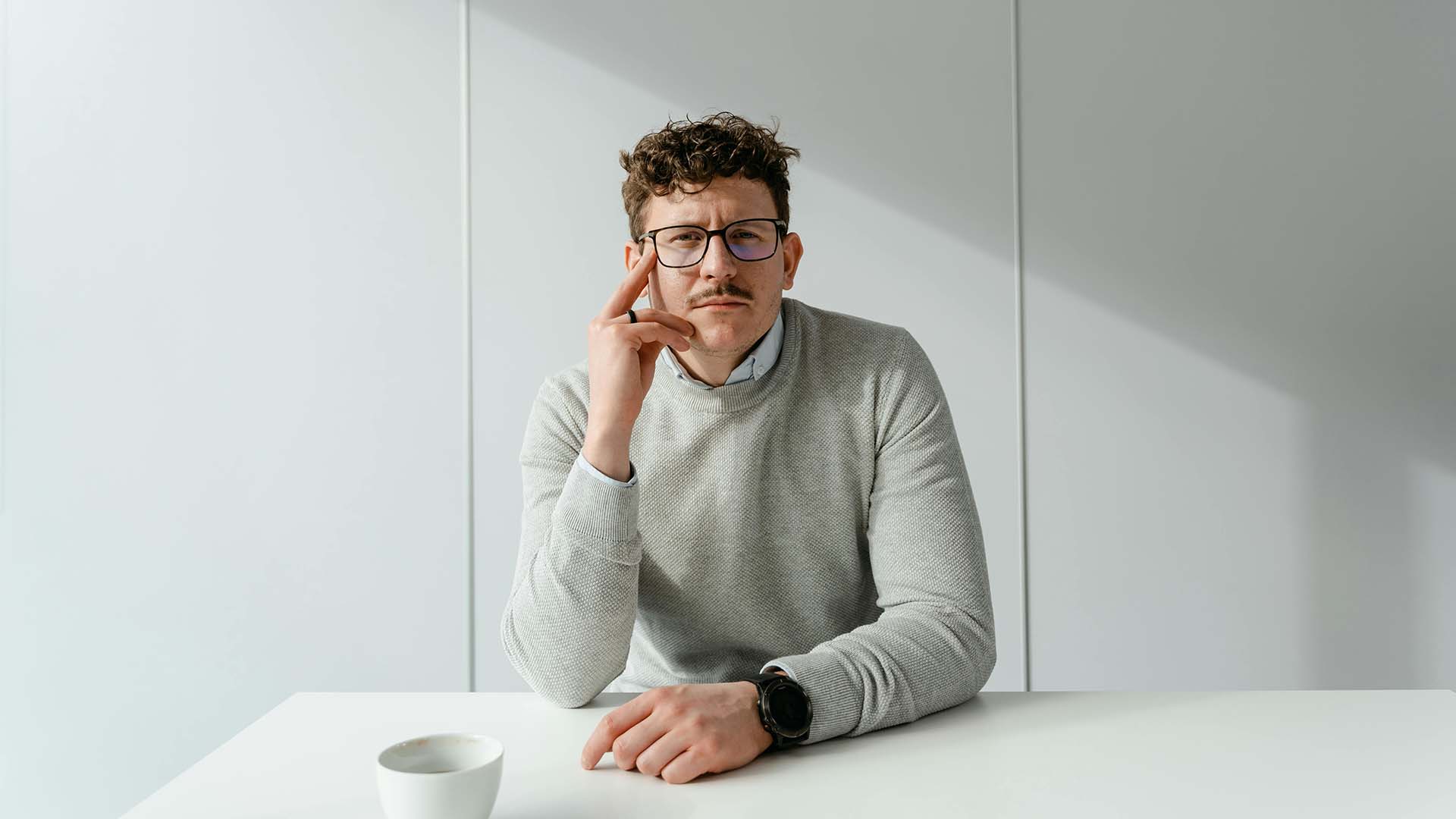 A man with curly hair and glasses sitting at a white desk with a coffee cup, resting his chin on his hand with a contemplative, exhausted expression