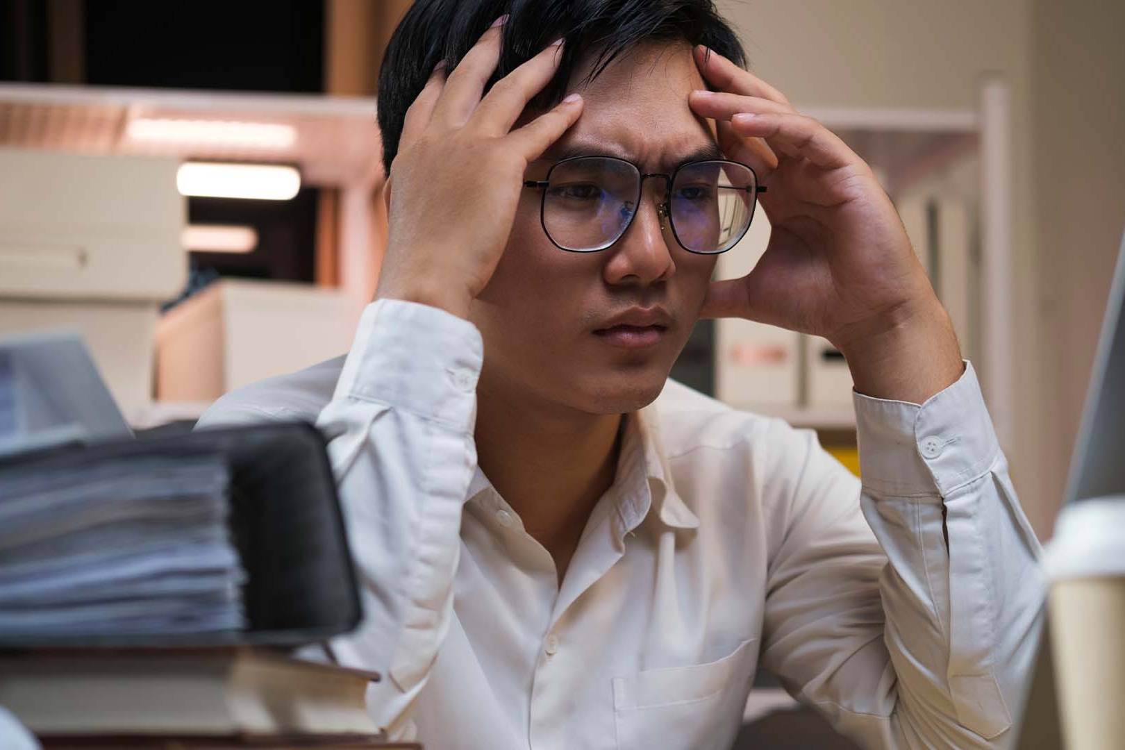 Stressed professional in shirt holding head at desk with laptop and papers in office