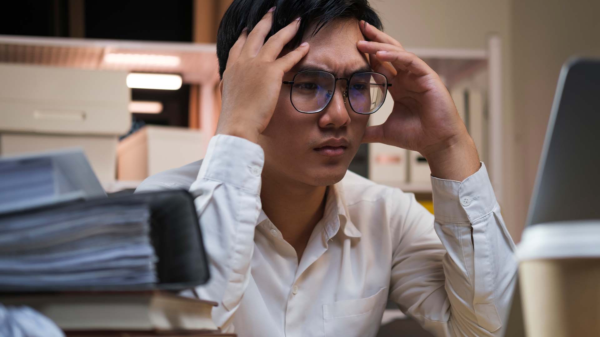 Stressed professional in shirt holding head at desk with laptop and papers in office