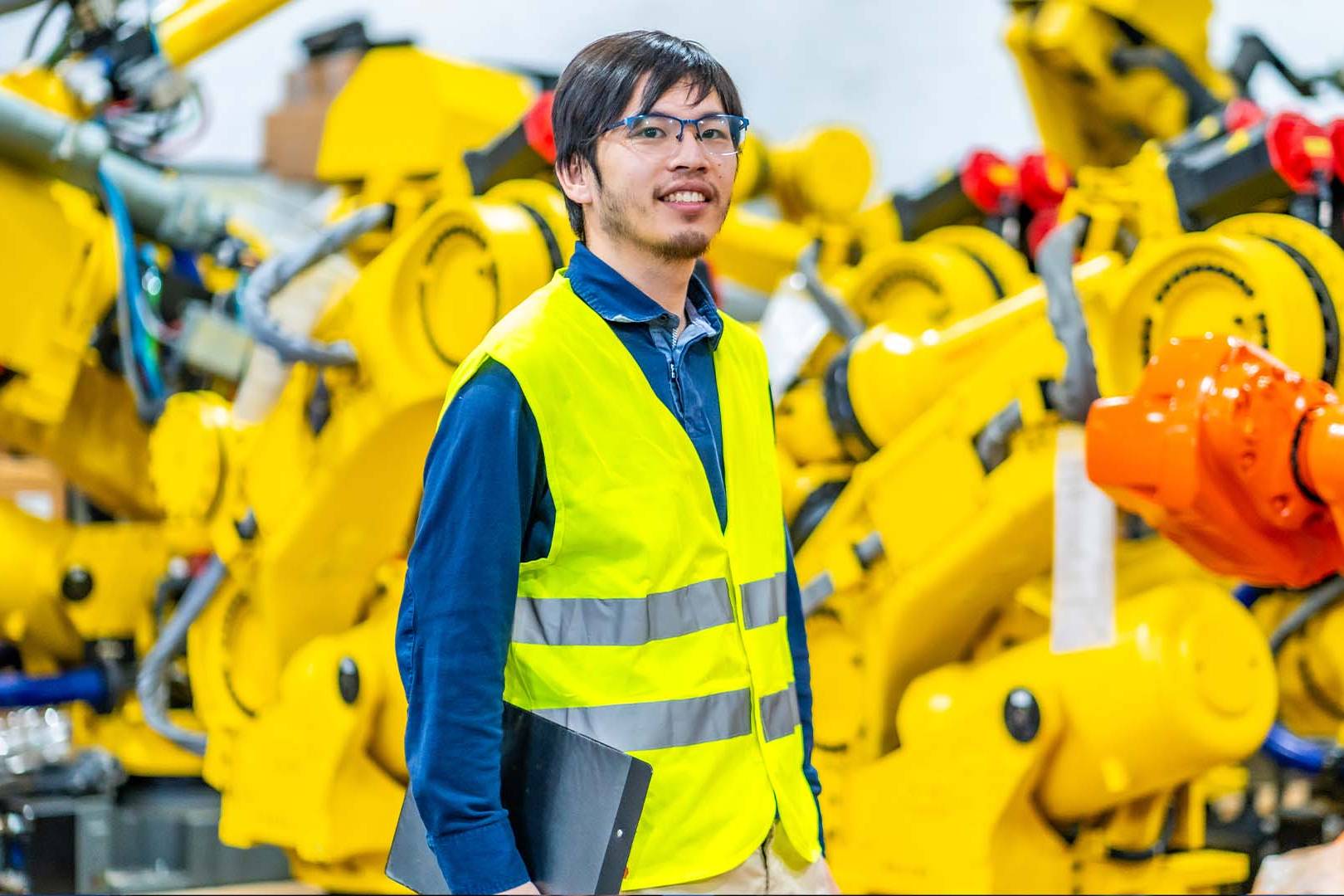 Robotics engineer in safety vest holding laptop standing among industrial robotic arms in tech manufacturing facility