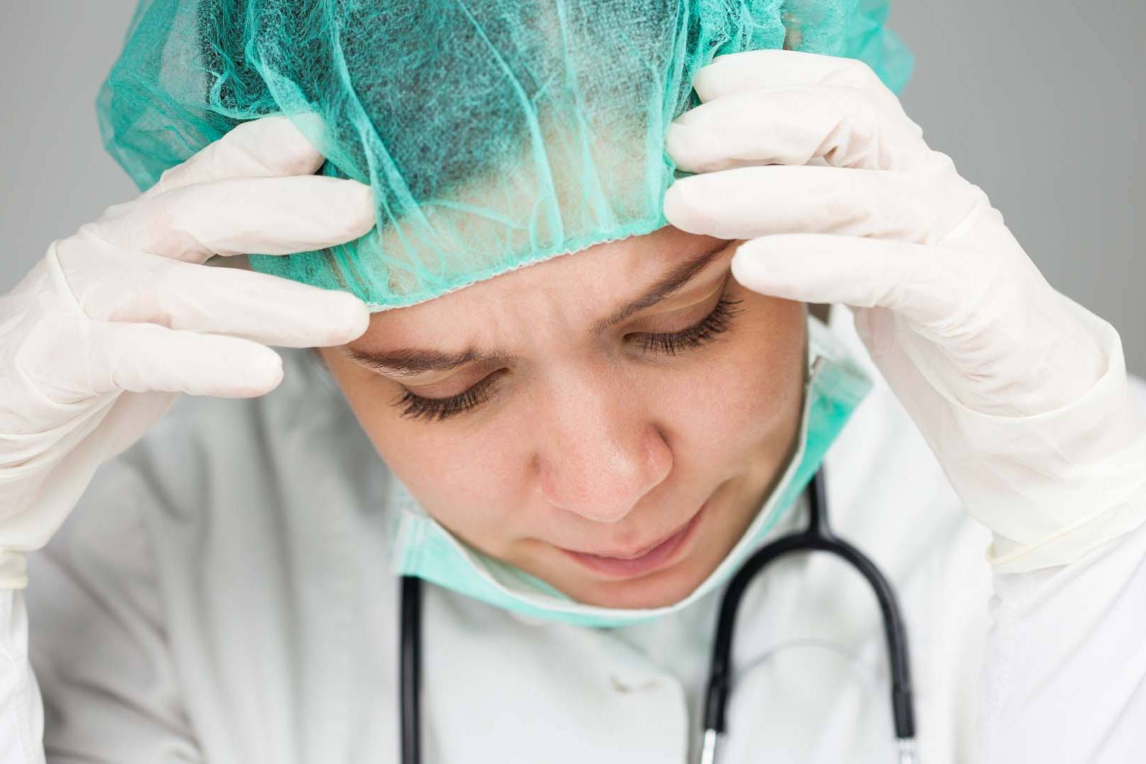 Stressed female doctor in surgical cap and gloves holding head in exhaustion