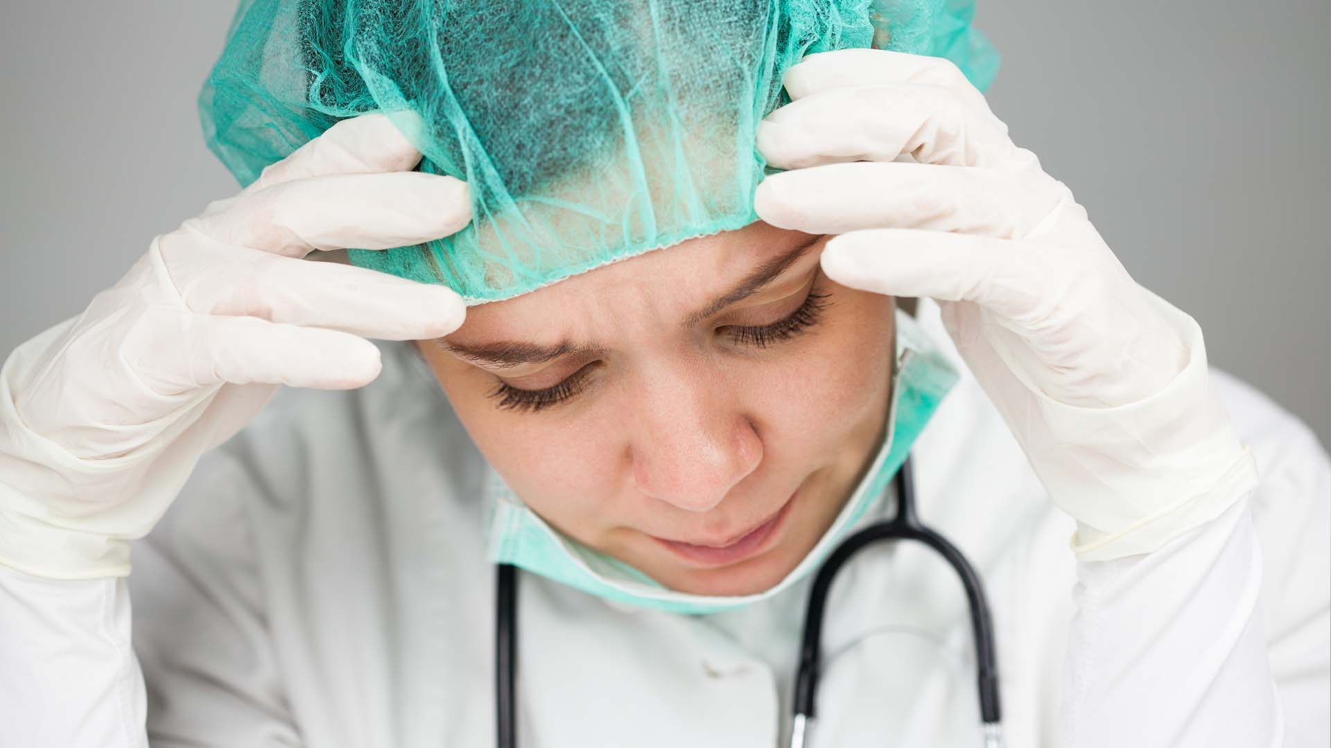 Stressed female doctor in surgical cap and gloves holding head in exhaustion
