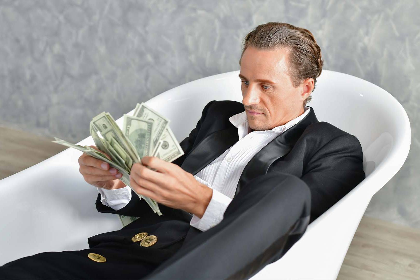 Sophisticated man in tuxedo counting cash while lounging in a bathtub