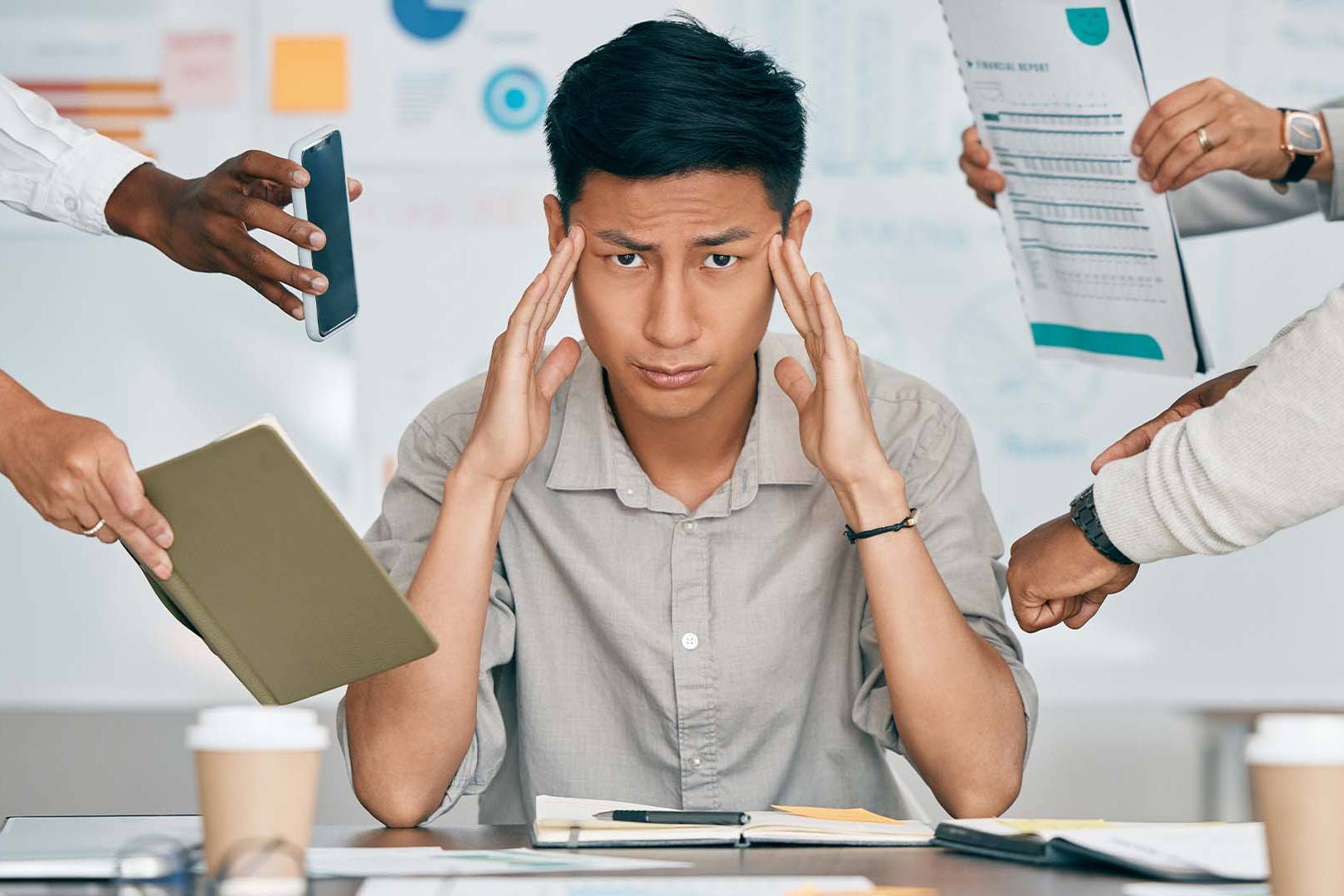 Overwhelmed executive holding head amid multiple hands offering documents in office