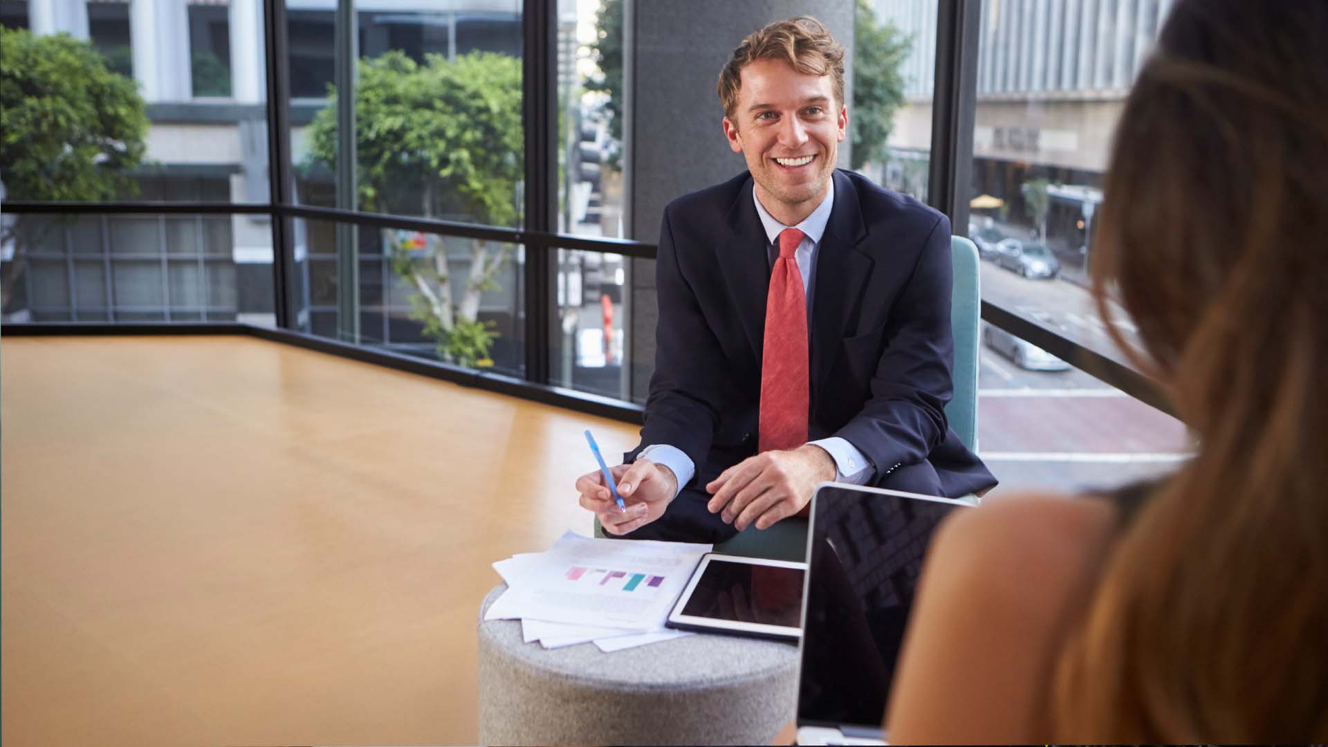 Investment banker in navy suit with red tie reviewing financial charts in downtown Los Angeles high-rise conference room