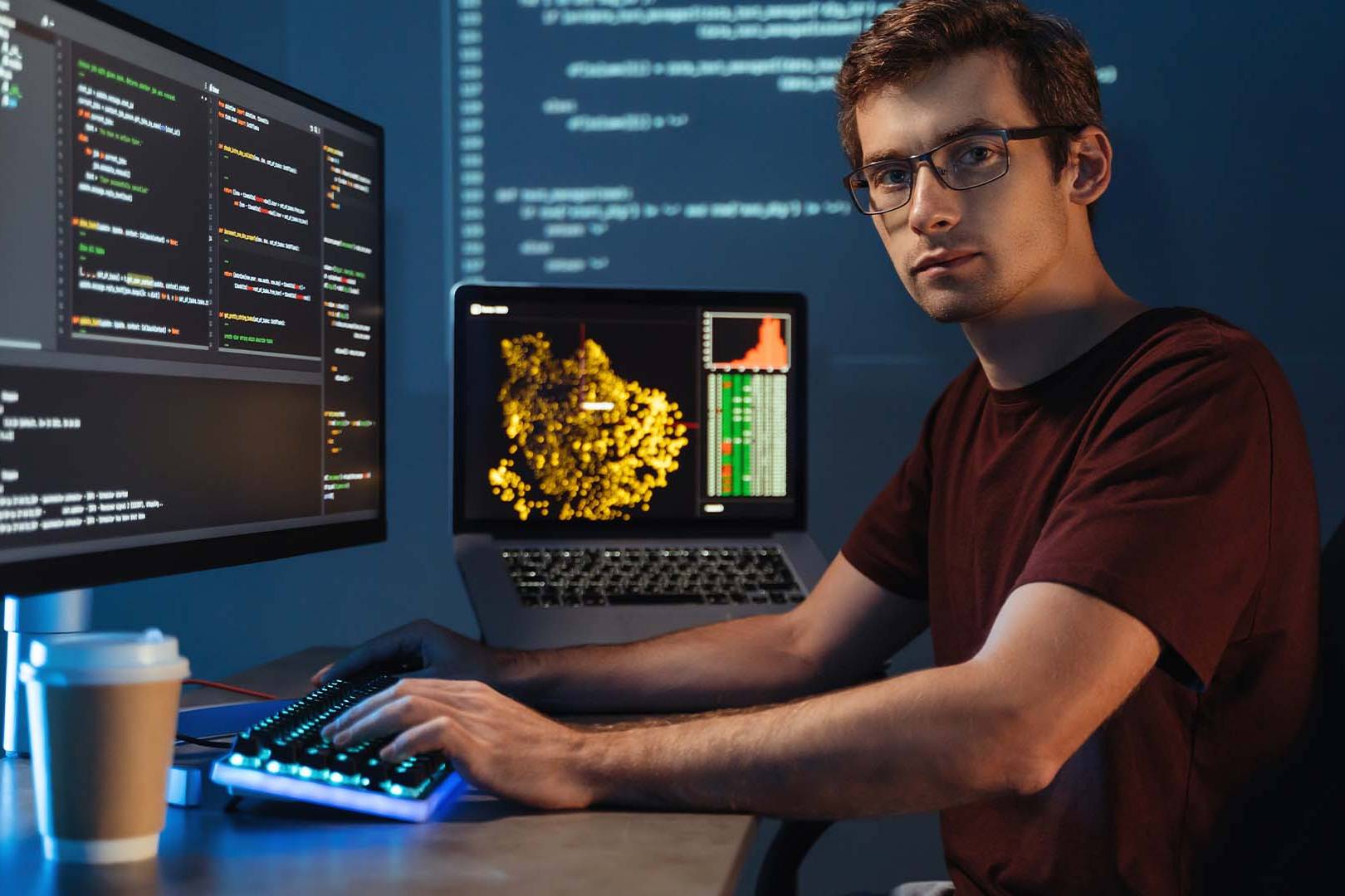 Focused programmer with glasses typing code on dual monitors in dimly lit workspace with coffee