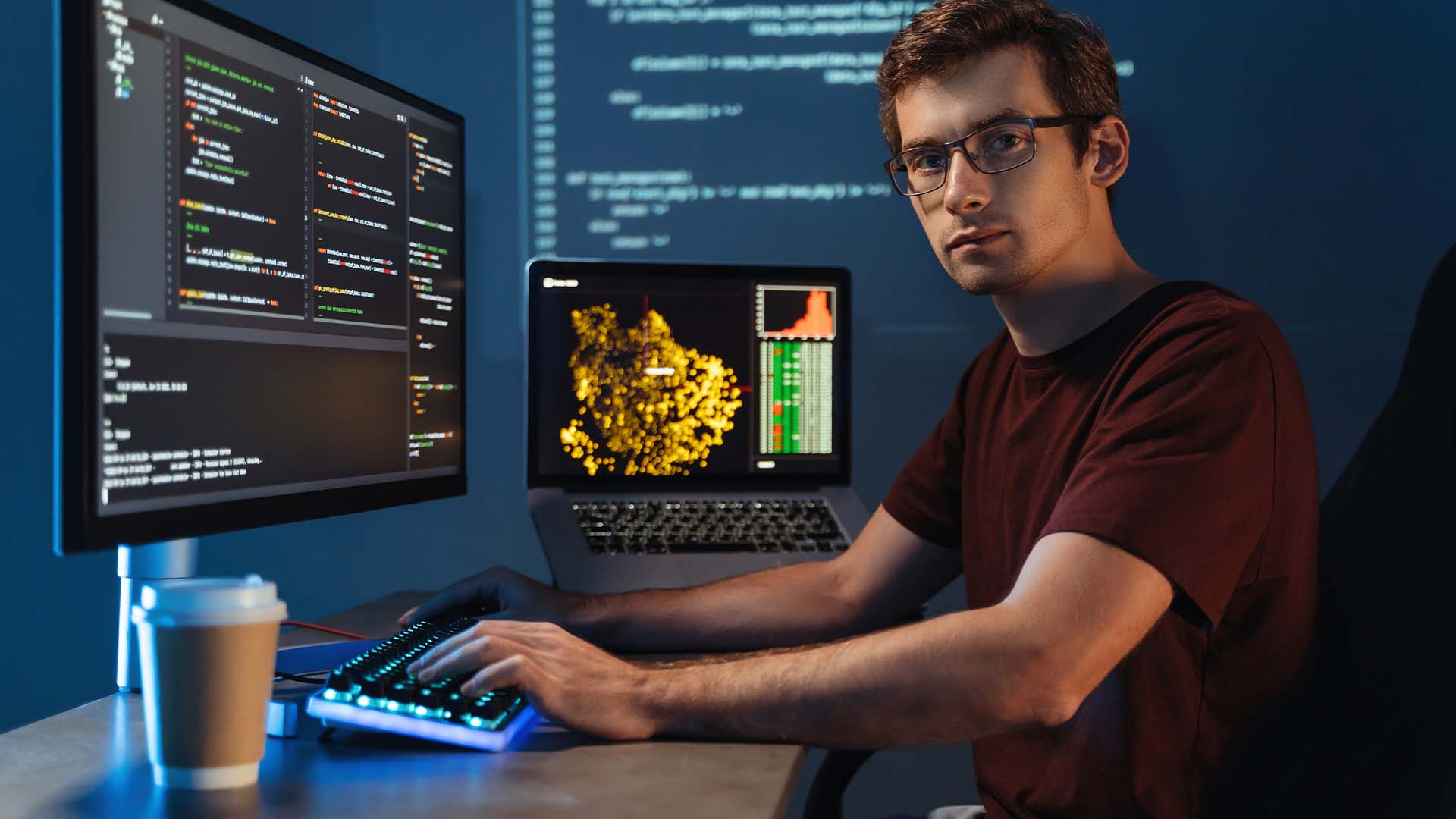 Focused programmer with glasses typing code on dual monitors in dimly lit workspace with coffee