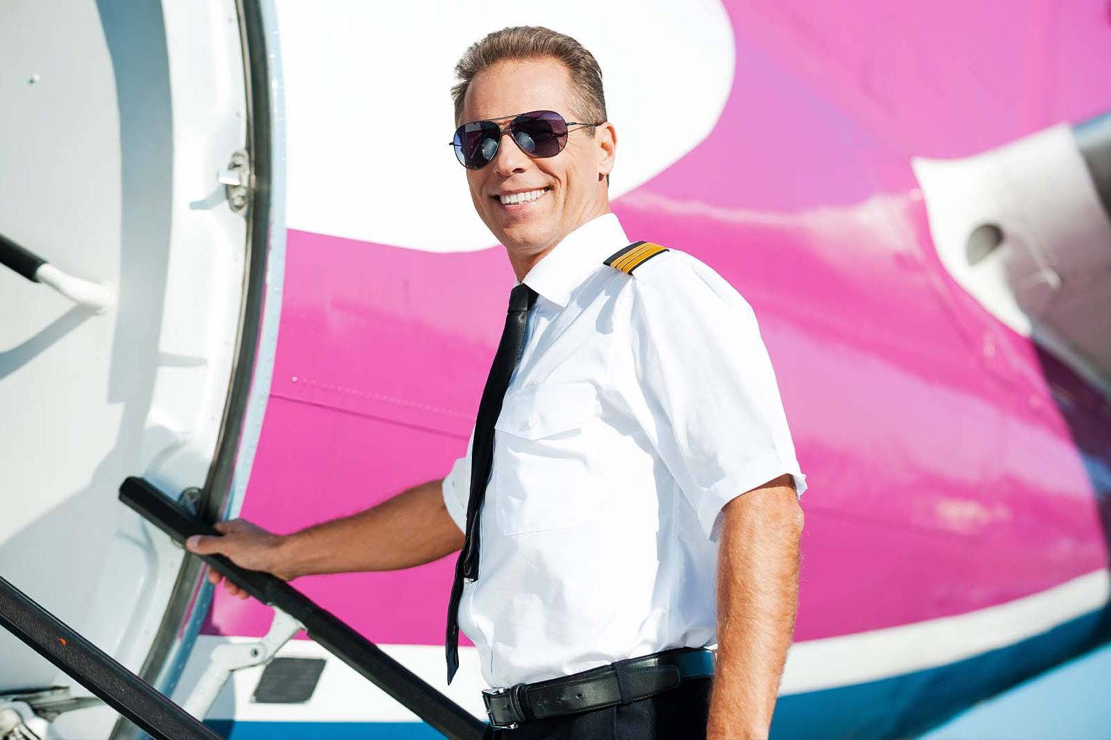 Confident airline pilot in uniform and sunglasses smiling while boarding colorful airplane