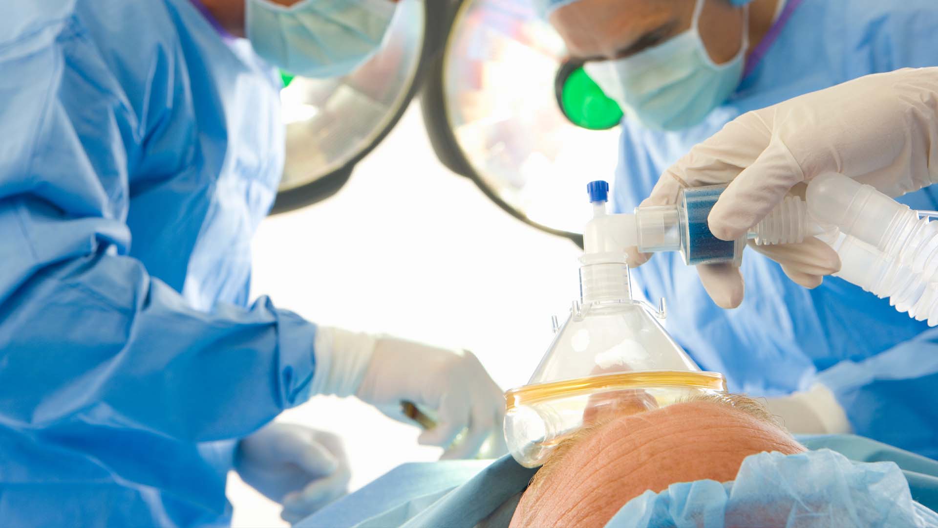 Gloved hands of anesthesiologist placing oxygen mask with breathing apparatus over patient's face during surgical procedure while medical team works in blue scrubs