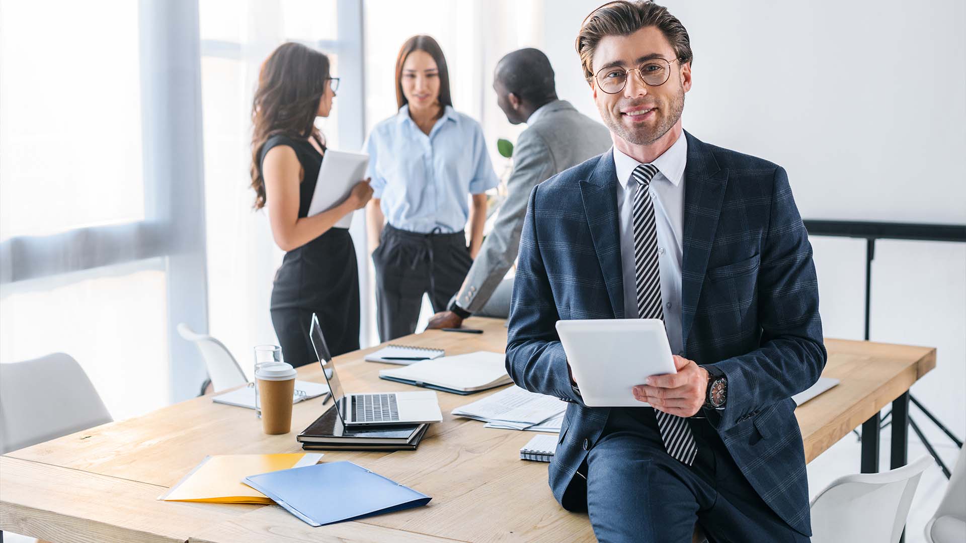 Smiling executive in suit holding tablet at office desk with diverse team in background