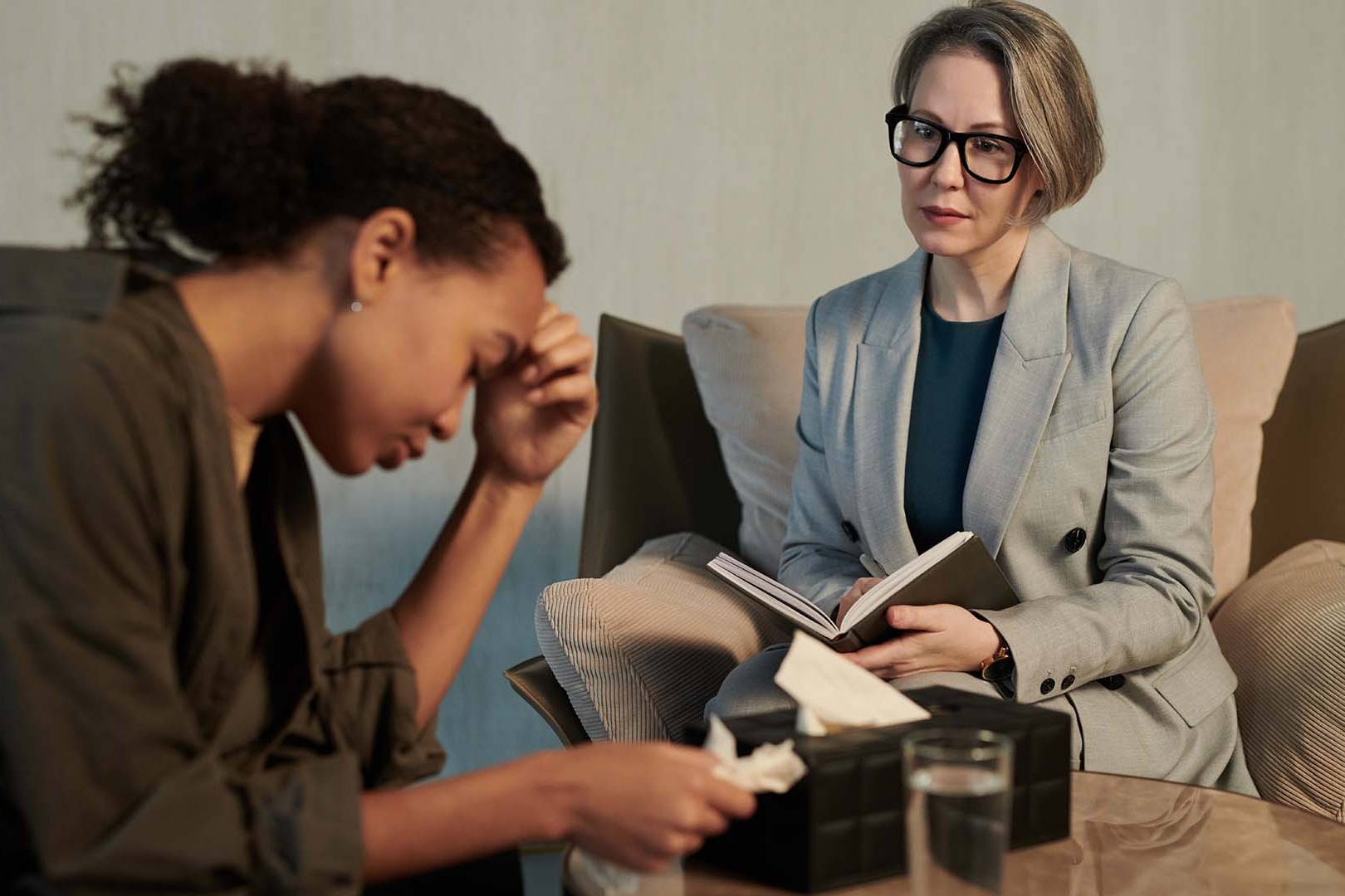 Female therapist in gray blazer and glasses holding notebook observing distressed male client in brown hoodie with head down during therapy session in modern office
