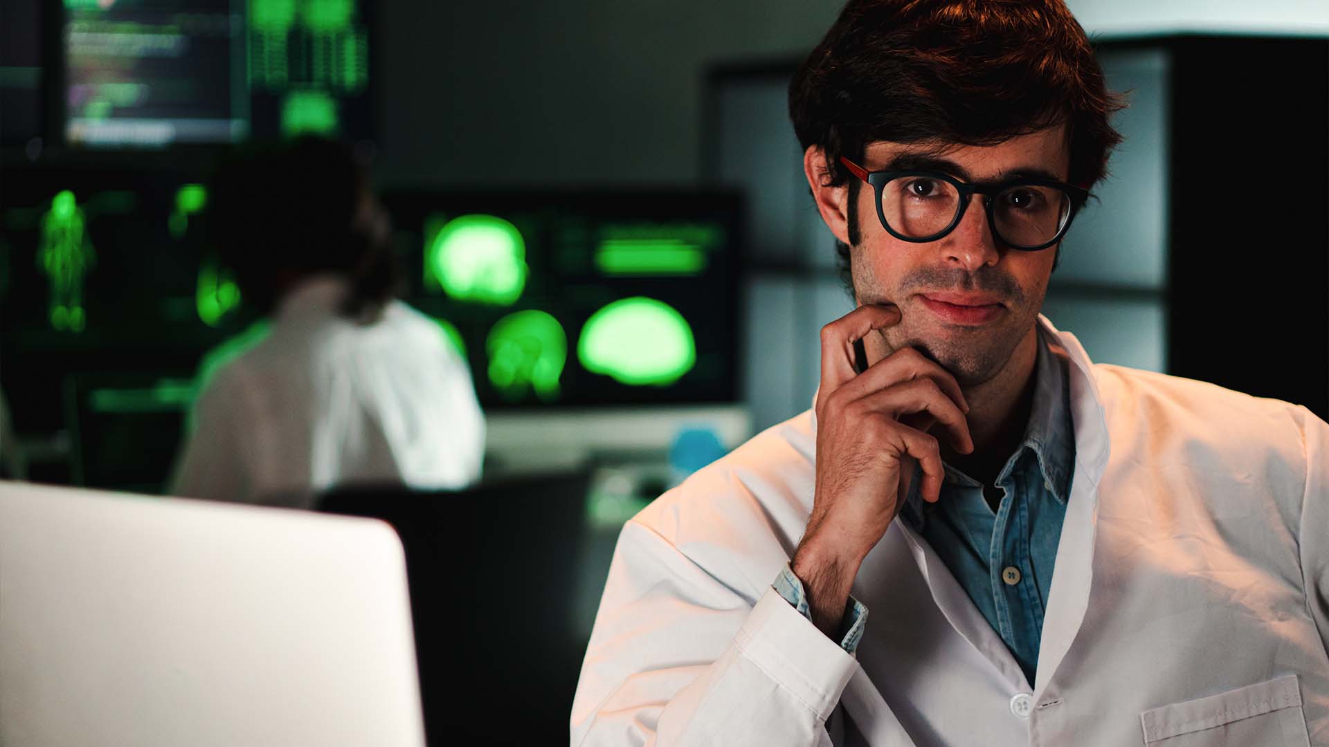 Young researcher in lab coat thoughtfully posing in a neuroscience lab with brain scans on monitors