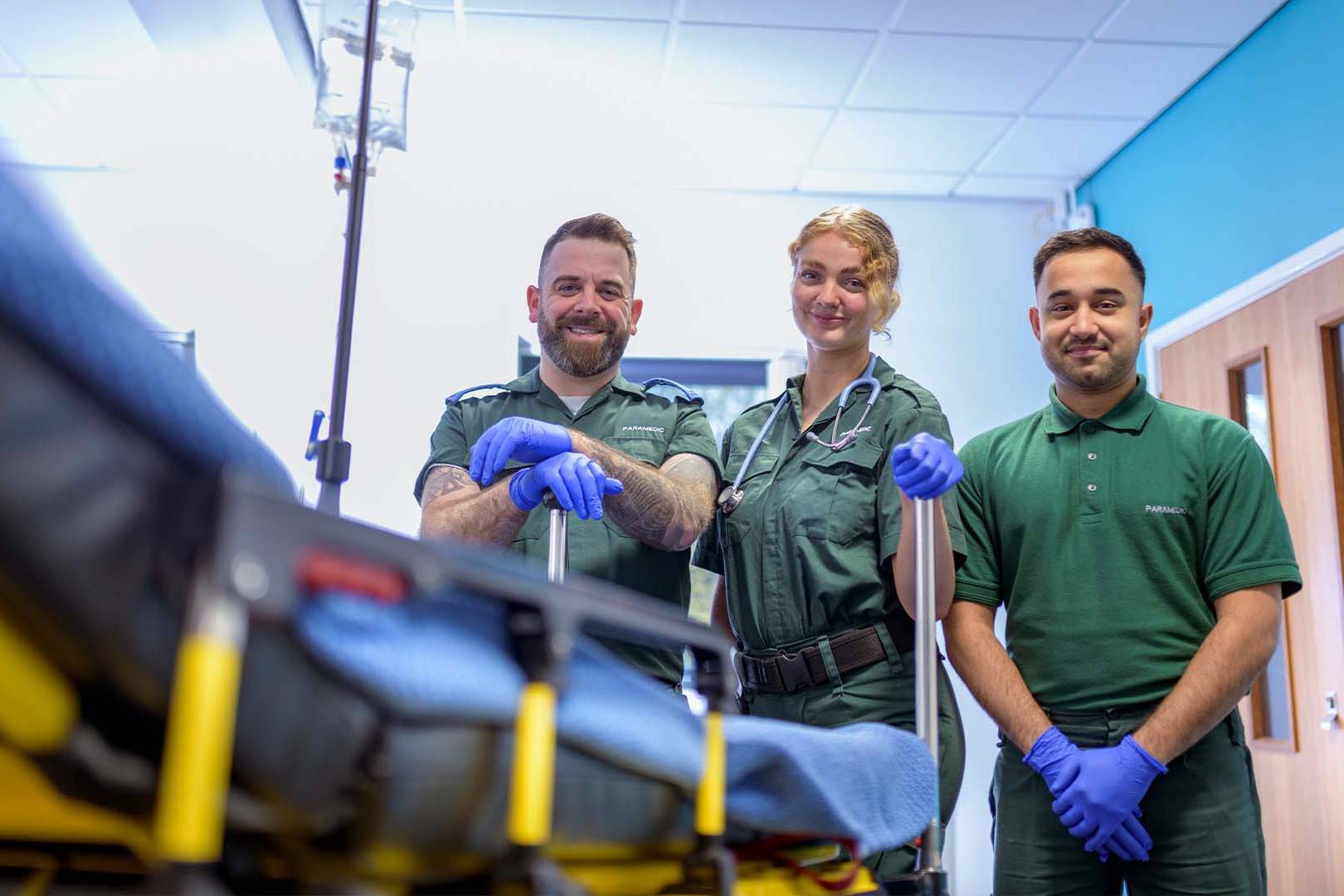 Three smiling paramedics in green uniforms and blue medical gloves standing confidently beside a stretcher with medical equipment in a bright modern emergency facility