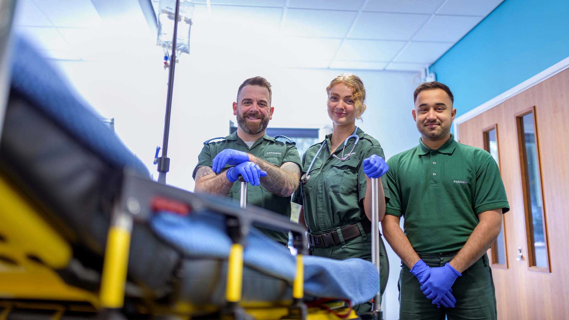 Three smiling paramedics in green uniforms and blue medical gloves standing confidently beside a stretcher with medical equipment in a bright modern emergency facility
