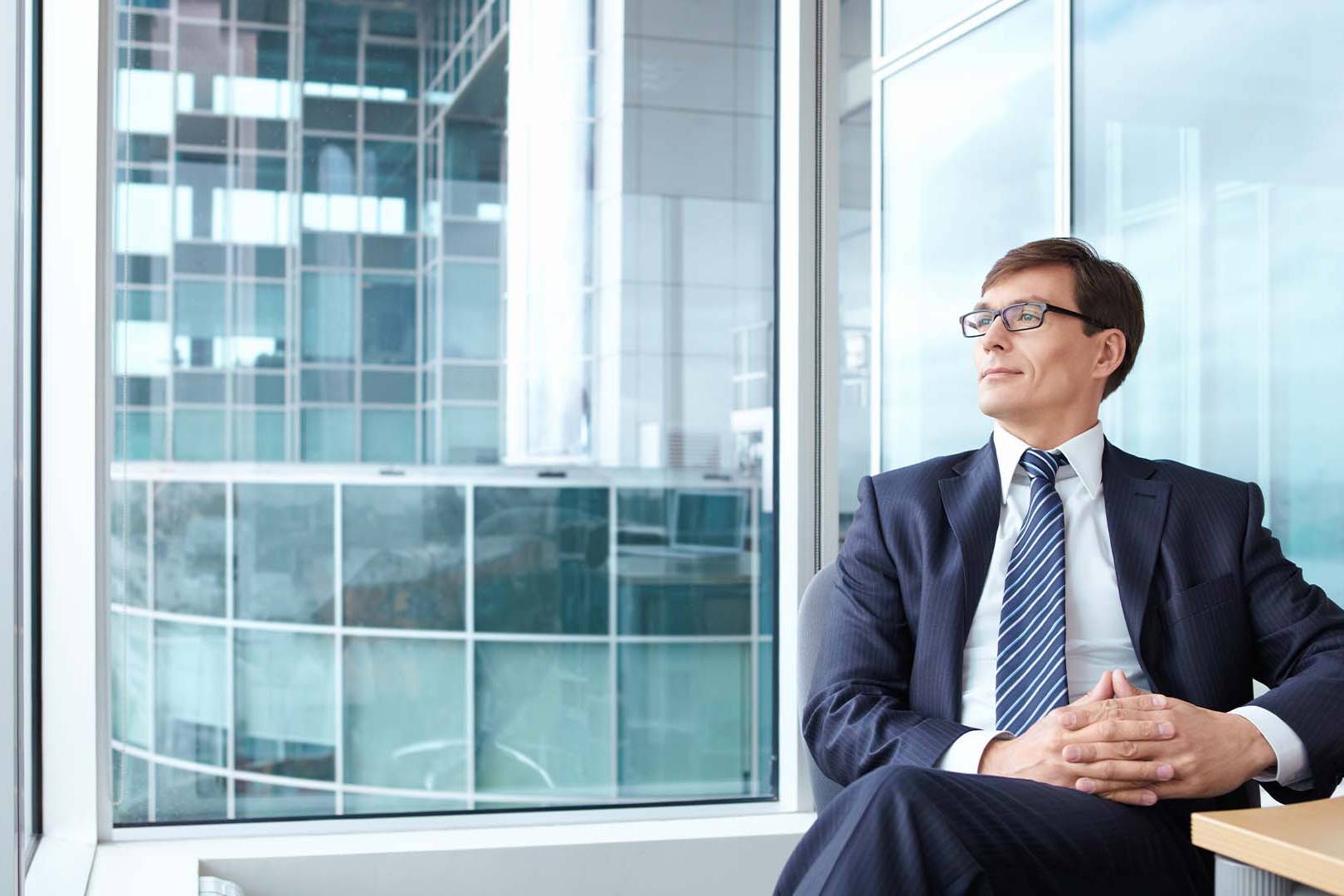 Exhausted executive in business suit gazing out corporate office window, representing the dangerous state of making critical decisions while experiencing burnout