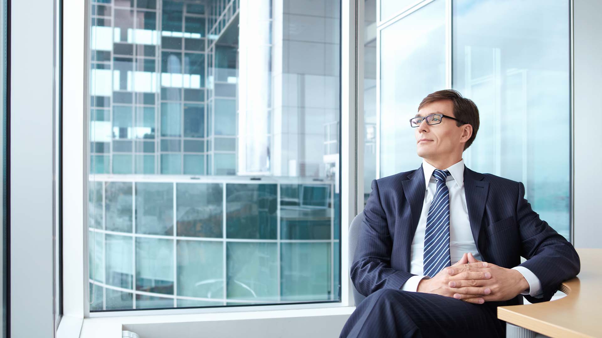 Exhausted executive in business suit gazing out corporate office window, representing the dangerous state of making critical decisions while experiencing burnout