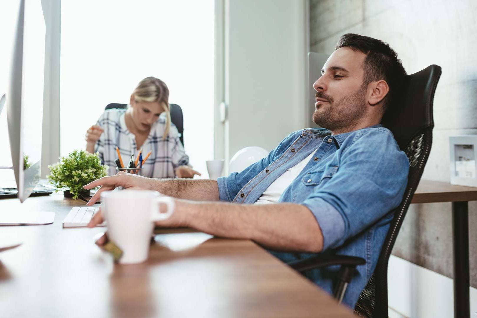 Exhausted startup founder slumped at desk with coffee while team member works in background, representing complete depletion from entrepreneurial demands