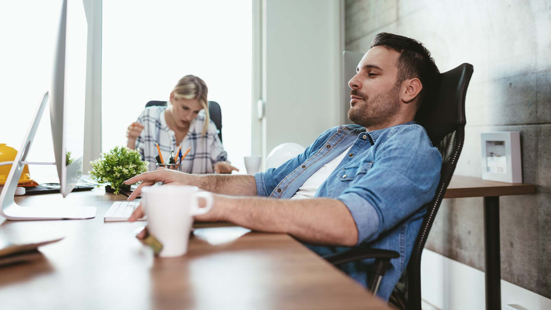 Exhausted startup founder slumped at desk with coffee while team member works in background, representing complete depletion from entrepreneurial demands