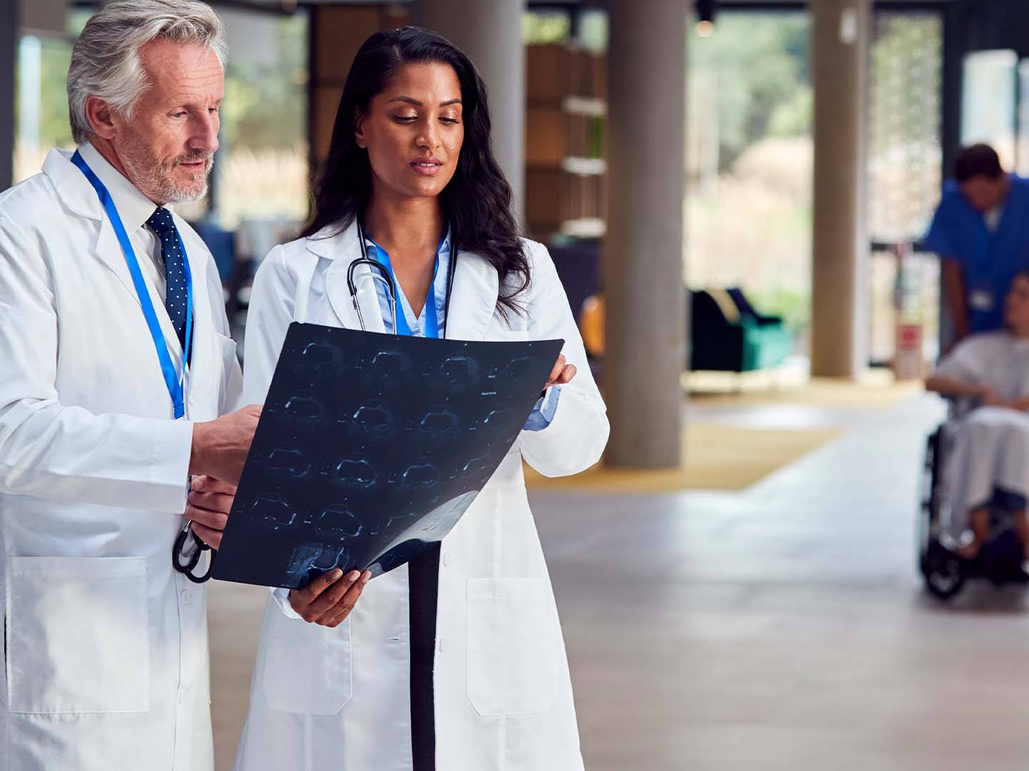 Two physicians in white coats reviewing medical scans in hospital corridor while patient care continues in background