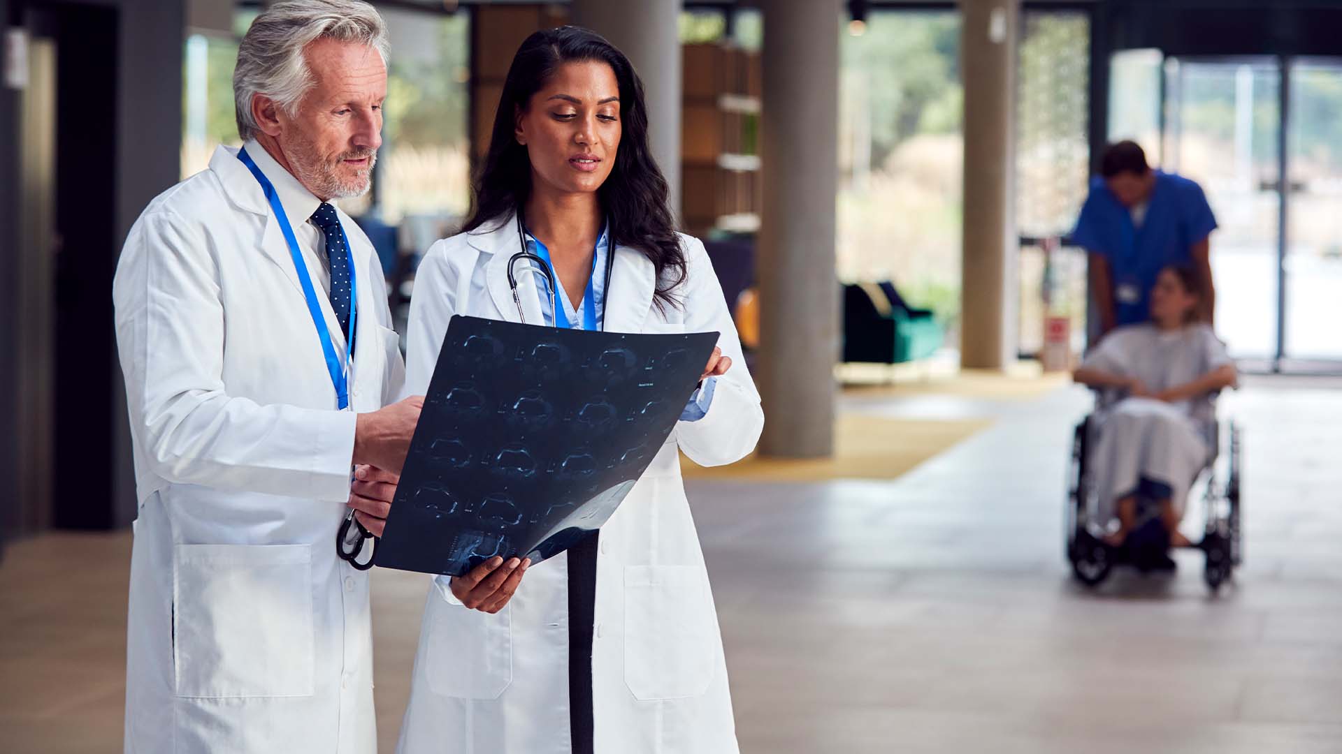 Two physicians in white coats reviewing medical scans in hospital corridor while patient care continues in background