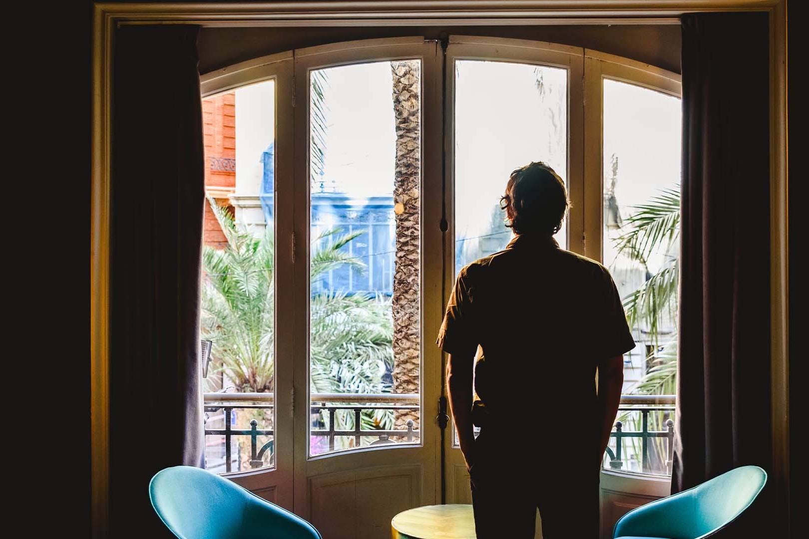 Person standing alone looking out window in contemplation, representing the relationship difficulties and emotional isolation experienced by high-achieving professionals