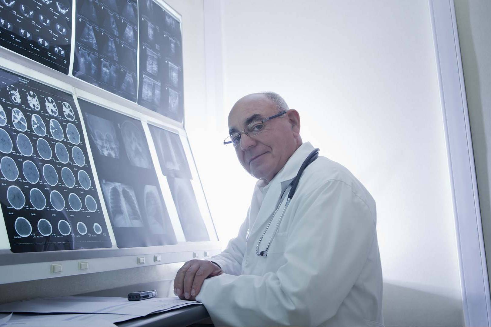 Male neurologist in white coat with glasses and stethoscope reviewing multiple brain CT and MRI scan images displayed on illuminated medical monitors in radiology room